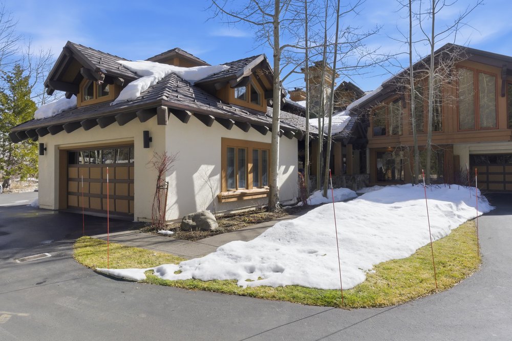 This is a front exterior view of a luxurious home featuring a combination of stucco and wood finishes. The house has a multi-car garage, multiple windows, and a complex roofline with snow accumulation. The front yard has patches of grass and snow, with a paved driveway leading up to the garages, creating an inviting and upscale curb appeal.
