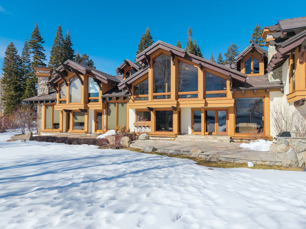 This is a front exterior view of a luxurious mountain home, showcasing its architectural design with large windows, wooden beams, and stone accents. The property is surrounded by snow-covered ground and tall evergreen trees, creating a serene and picturesque setting. The home's design emphasizes natural light and blends seamlessly with the surrounding landscape.