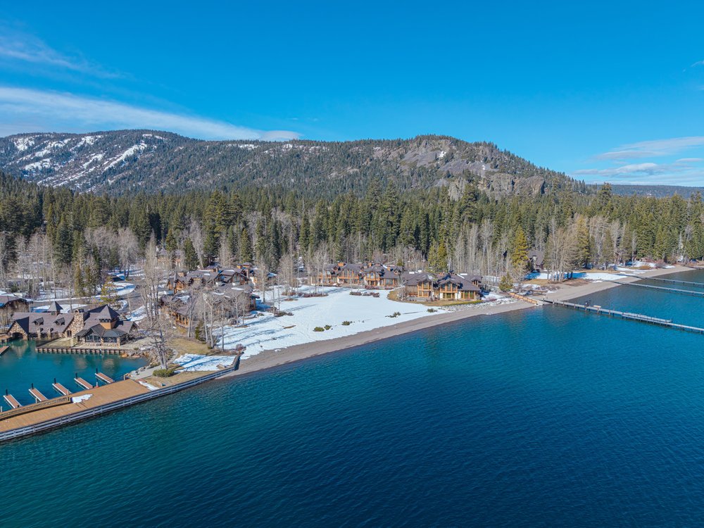 This aerial view showcases a luxurious lakeside property with multiple residences nestled among trees and a snow-dusted landscape. A long pier extends into the clear blue water, and the architecture features a blend of rustic and modern elements. The scene conveys a sense of exclusivity and natural beauty, ideal for high-end real estate.