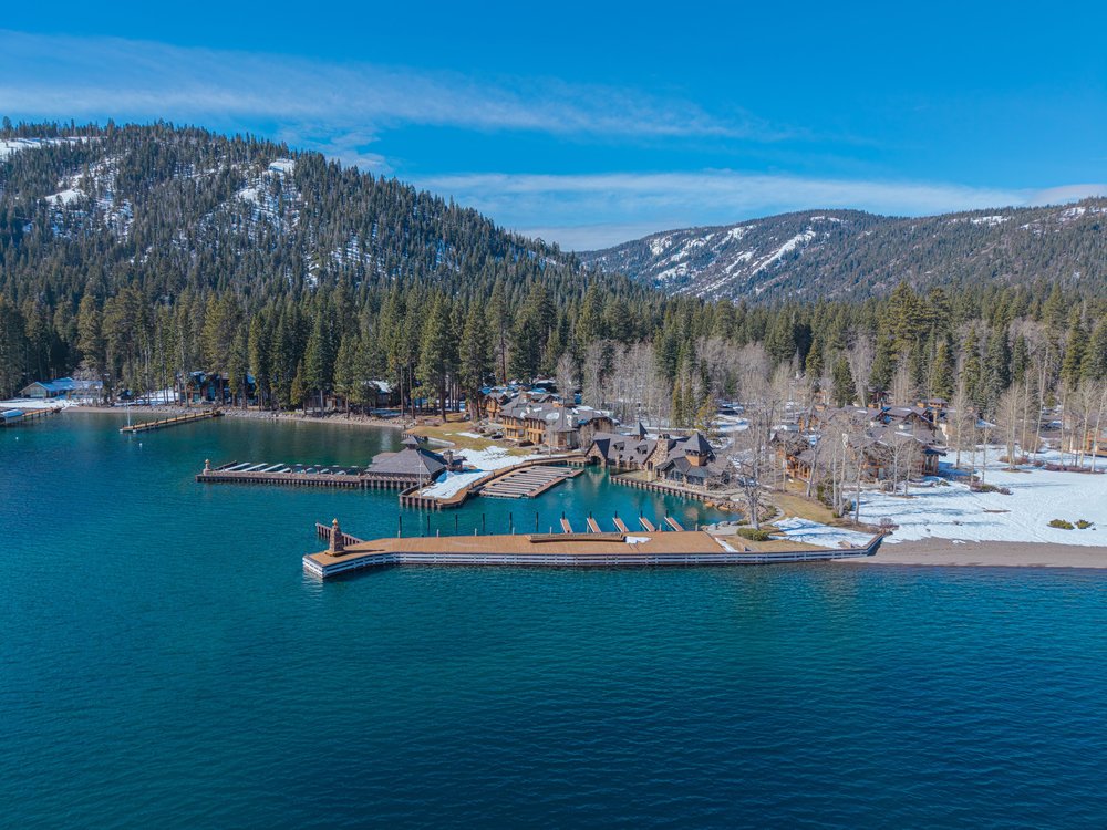 This aerial shot showcases a stunning lakeside property with multiple docks extending into the clear blue water. The property features several buildings with unique architectural designs, nestled among lush evergreen trees and patches of snow. Mountains rise in the background, creating a picturesque and serene setting.