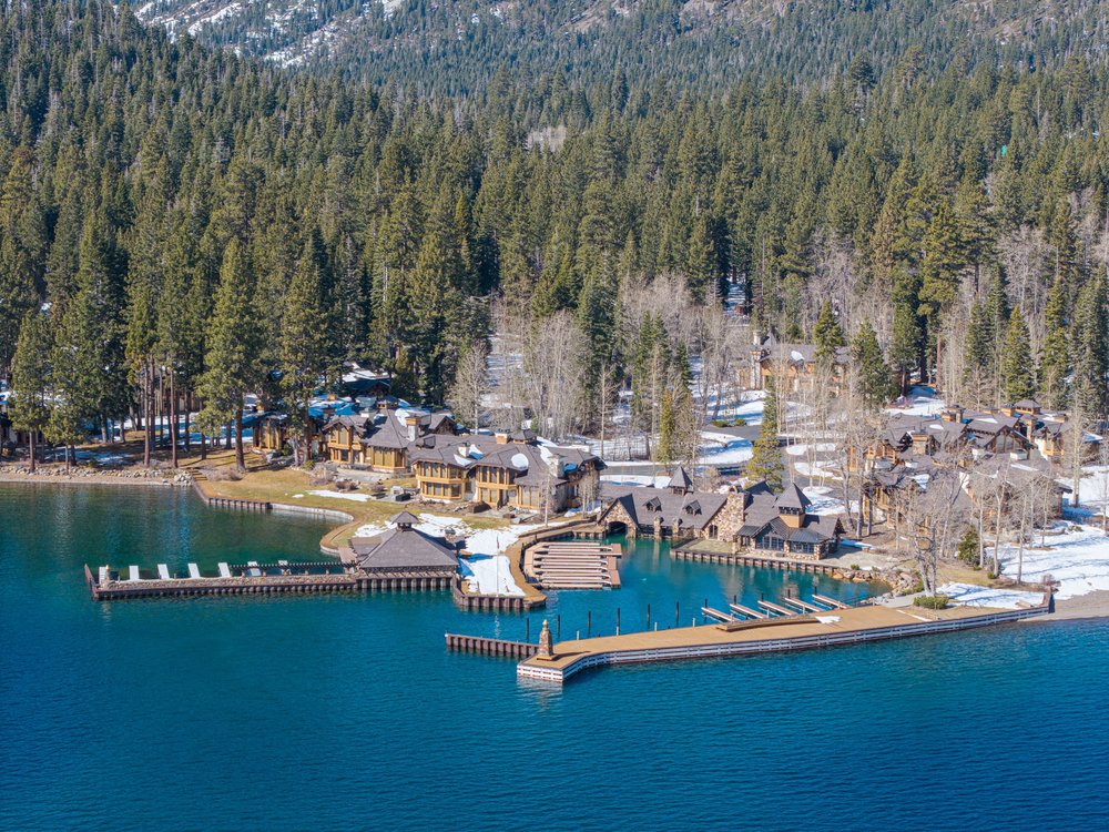 This aerial view showcases a luxurious lakeside estate with multiple buildings, extensive docks, and boat slips. The architecture features natural stone and wood, blending seamlessly with the surrounding forest. The clear blue water and snow-dusted landscape create a serene and exclusive atmosphere.