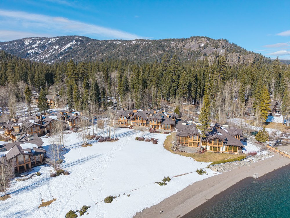 This aerial view showcases a collection of luxurious homes nestled amidst a snowy landscape and dense forest, with a mountain range in the background. The properties feature architectural designs with wood and stone elements, complemented by large windows that offer stunning views. The scene captures a serene and upscale residential setting, ideal for those seeking privacy and natural beauty.