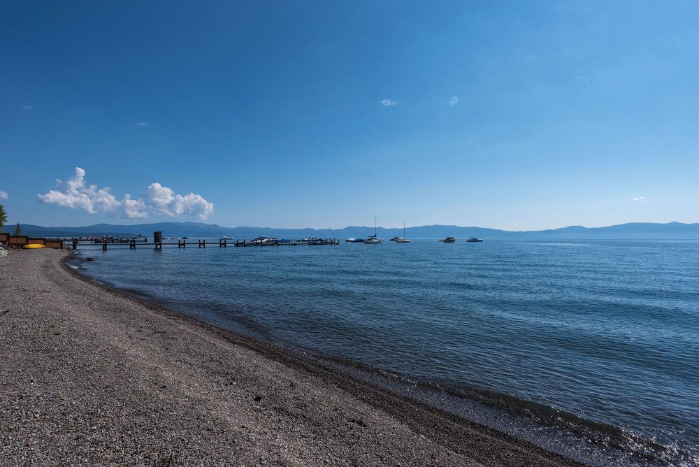 The image showcases a serene beachfront property with a pebble beach leading to calm, blue waters under a clear sky. A wooden pier extends into the water, dotted with boats and offering a picturesque view of the distant mountains. The scene evokes a sense of tranquility and natural beauty, ideal for a waterfront real estate listing.