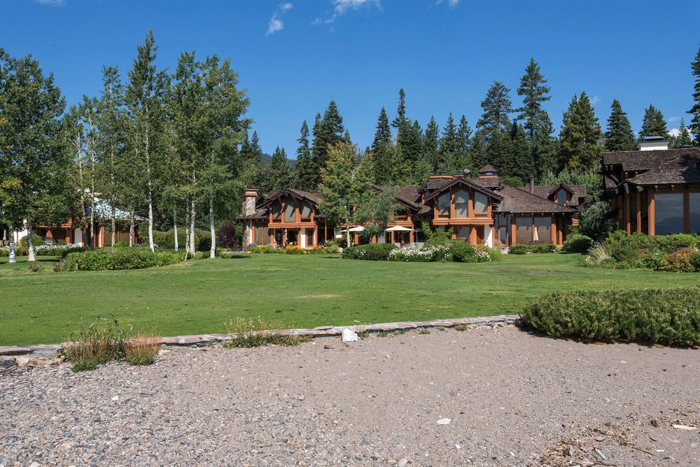 This image showcases a sprawling yard with a well-manicured lawn leading up to a luxurious home. The property features mature trees, landscaping, and a gravel shoreline, creating a serene and private setting. The architectural style of the home is rustic and elegant, blending seamlessly with the natural surroundings.