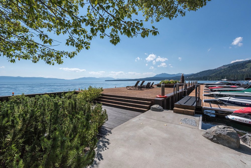 This image showcases a beautiful waterfront deck and dock area. The deck features wooden flooring and steps leading down to a concrete patio. Several boats are docked alongside the pier, and lounge chairs are arranged on the deck, creating an inviting space for relaxation and entertainment by the water.