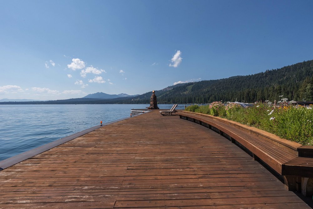 This image showcases a spacious wooden deck extending over a serene lake. The deck features built-in seating, lounge chairs, and a decorative stone structure, creating an inviting outdoor living space. The backdrop includes lush greenery and mountains, enhancing the property's appeal for relaxation and entertainment.