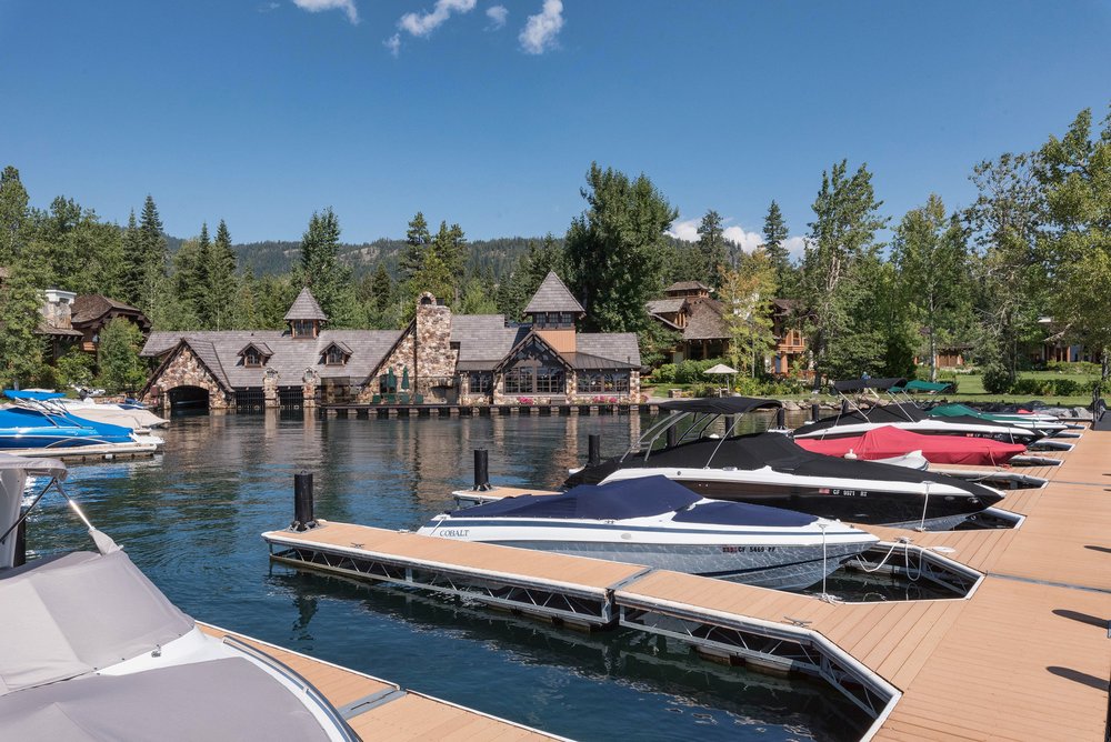 This exterior shot showcases a stunning boathouse with stone and wood architecture, nestled on the water's edge. Several boats are docked along the pier, adding to the property's appeal for water enthusiasts. The surrounding lush greenery and clear blue sky create a serene and luxurious atmosphere.