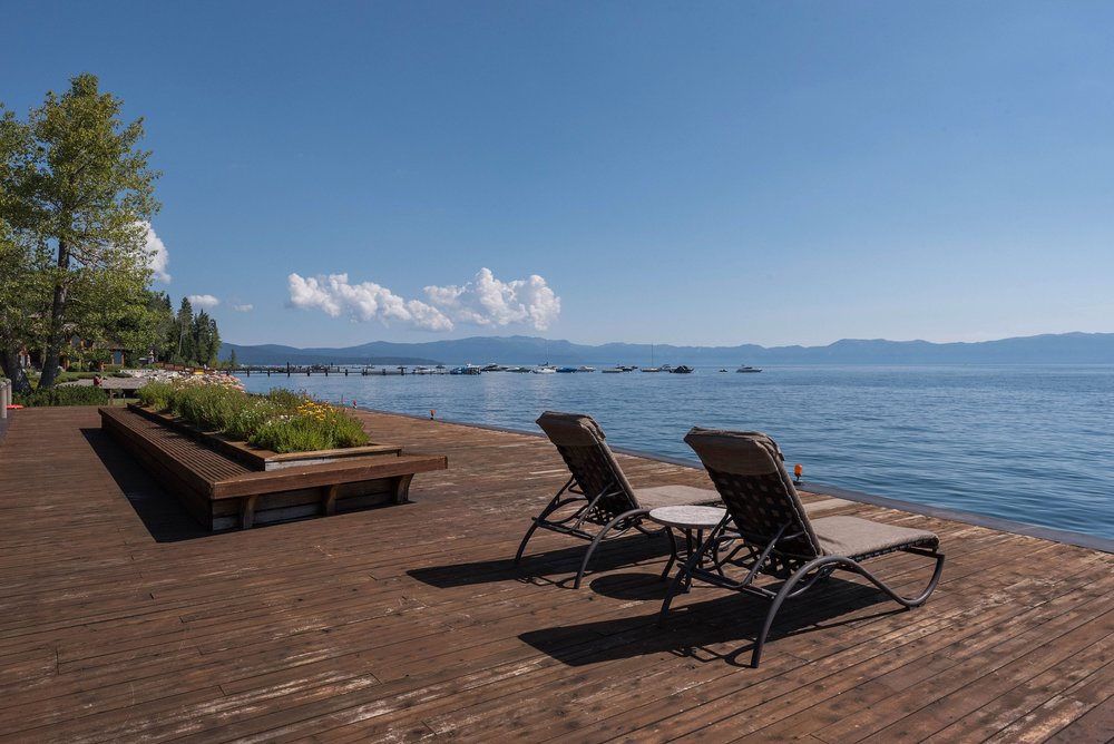 This image showcases a serene outdoor deck area with two lounge chairs and a small table, positioned to overlook a tranquil lake. The wooden deck features a built-in planter with greenery, adding a touch of nature to the space. The scene evokes a sense of relaxation and offers a picturesque view, ideal for enjoying the waterfront.