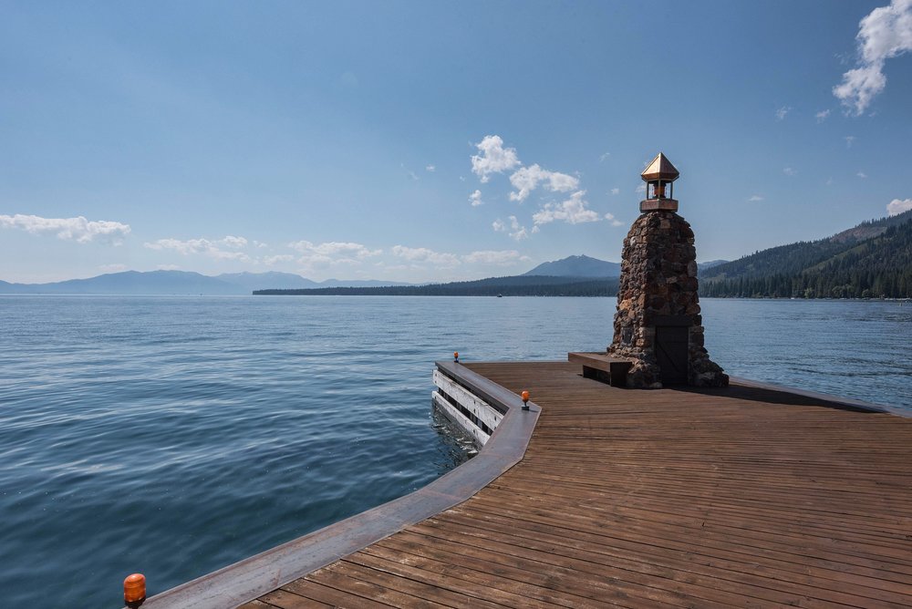 This image showcases a wooden deck extending over a serene body of water, possibly a lake or ocean. A stone structure with a decorative top sits prominently on the deck, adding a unique architectural element. The scene is bathed in natural light under a partly cloudy sky, creating a tranquil and inviting atmosphere.