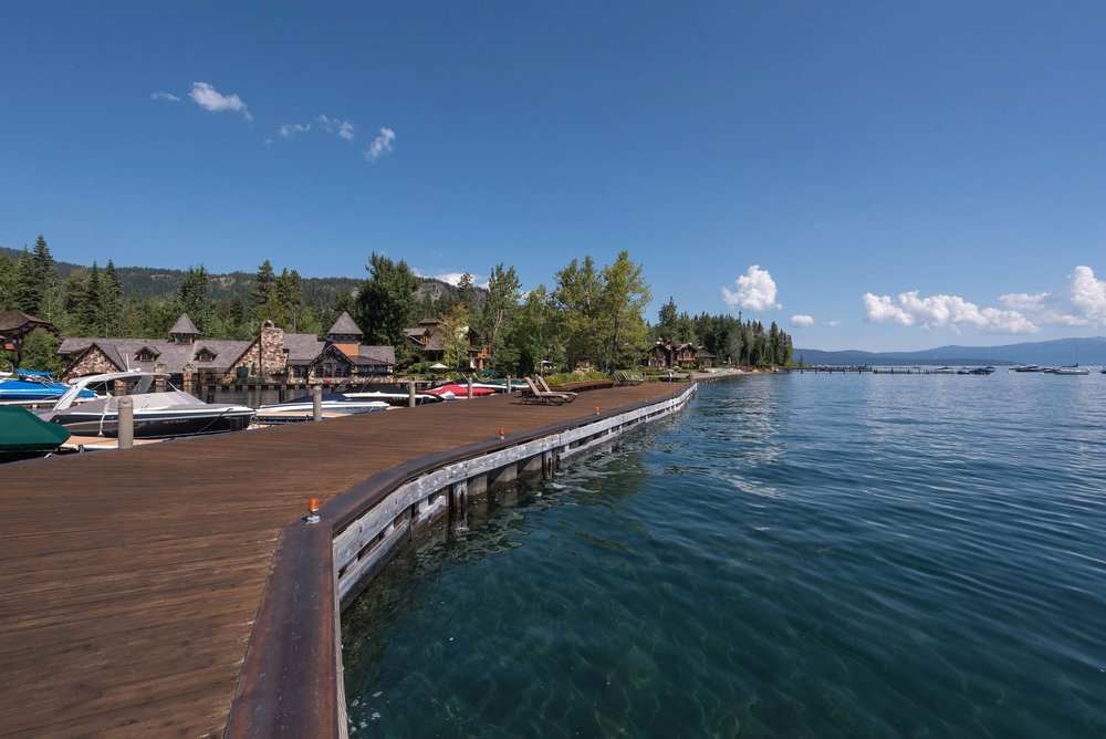 This image showcases a beautiful waterfront property with a large wooden deck extending over the water. The deck features railings and is lined with boats, offering direct access to the lake. In the background, there are luxurious homes nestled among trees, creating a serene and upscale atmosphere.