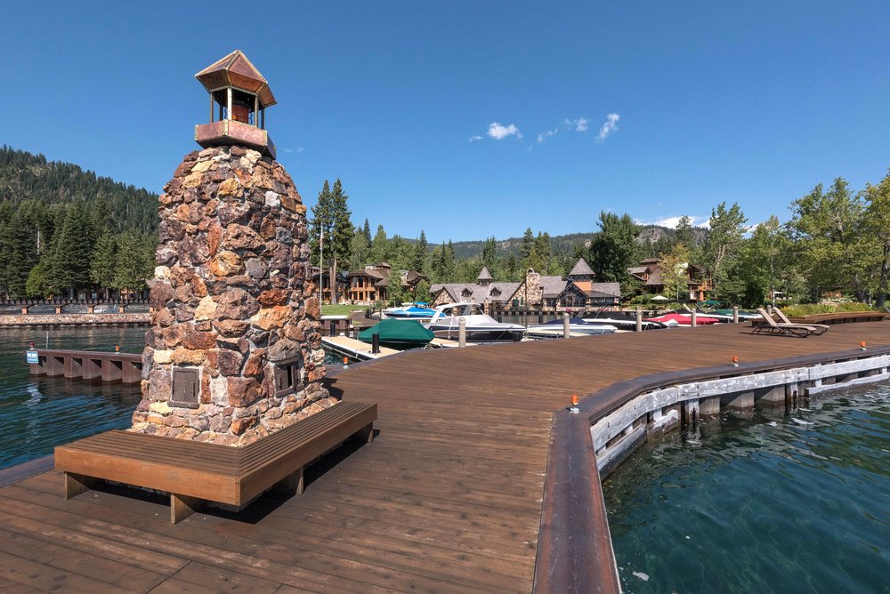 This image showcases a beautiful wooden deck extending over the water, featuring a unique stone structure with a wooden top and a built-in bench. The deck provides ample space for relaxation and entertainment, with boats docked nearby and luxurious homes visible in the background. The clear blue sky and calm water create a serene and inviting atmosphere.