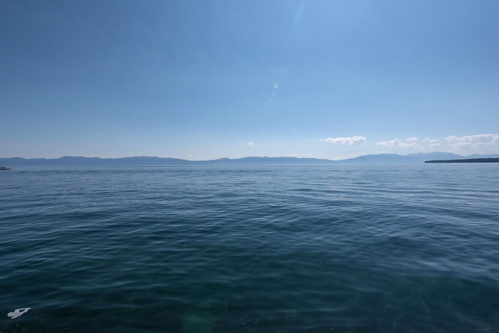 The image showcases a serene water view with a clear blue sky above. Gentle waves ripple across the water's surface, leading towards a distant shoreline with mountains. The overall impression is one of tranquility and natural beauty, suggesting a waterfront property or a location with scenic water access.