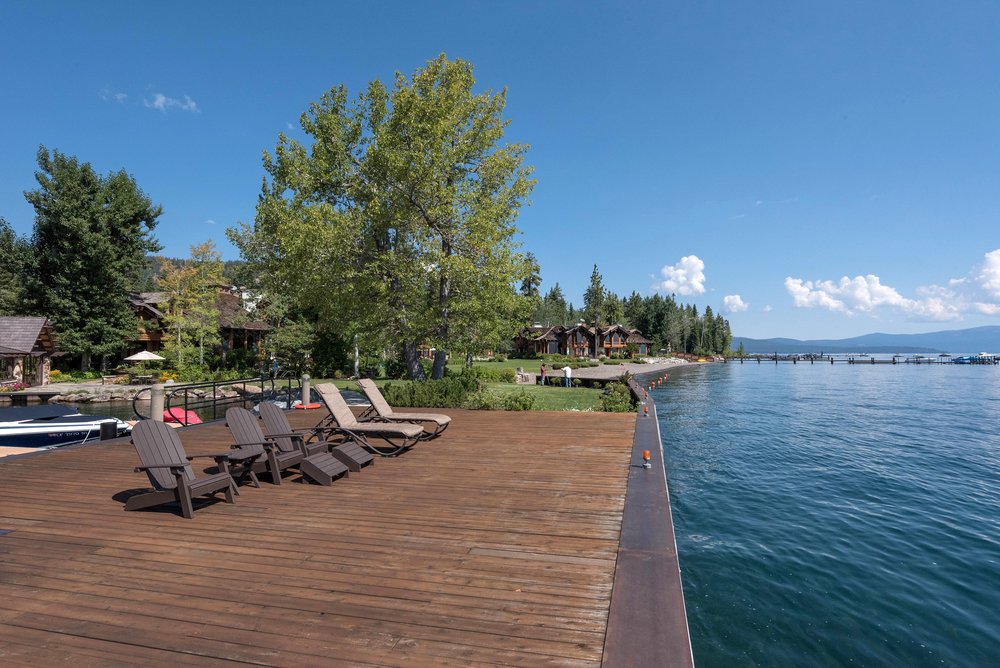 This image showcases a spacious wooden deck extending over the water, furnished with several Adirondack chairs and lounge chairs, creating an inviting outdoor living space. The deck offers stunning views of the lake and surrounding landscape, with a clear blue sky overhead. The scene evokes a sense of relaxation and luxury, perfect for enjoying waterfront living.