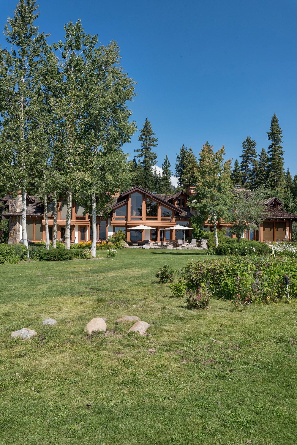 This image showcases the expansive yard and garden of a luxurious home. The well-manicured lawn is dotted with natural rock formations and leads up to a grand residence with large windows and a wood exterior. Mature trees frame the property, creating a sense of privacy and tranquility under a clear blue sky.