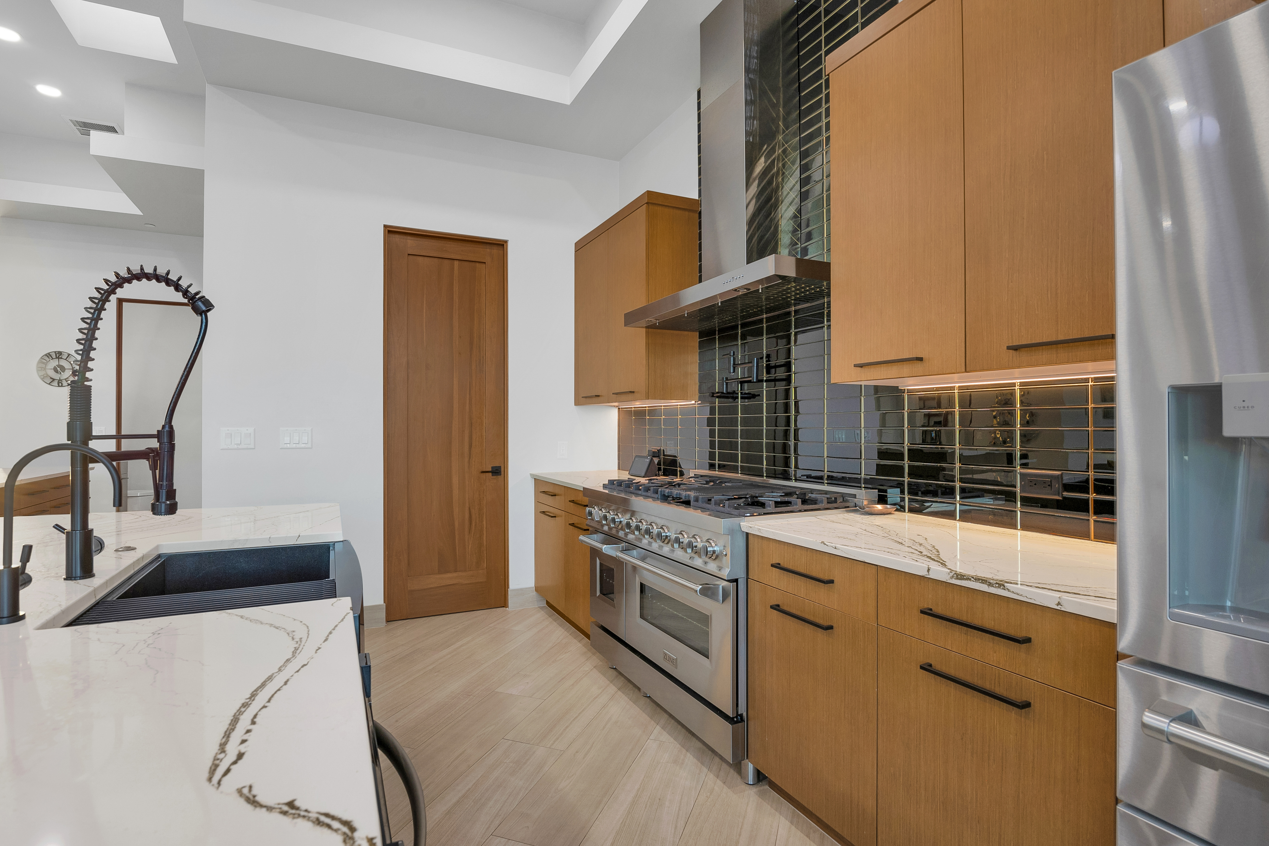 This modern kitchen features warm-toned wood cabinetry paired with sleek white stone countertops and a striking dark, reflective tile backsplash. A professional-grade stainless steel range with a matching hood serves as the focal point, while a large island with a dark sink and industrial-style faucet provides additional workspace. The space is finished with light-colored wood-look flooring and a clean, minimalist aesthetic that emphasizes high-end functionality.