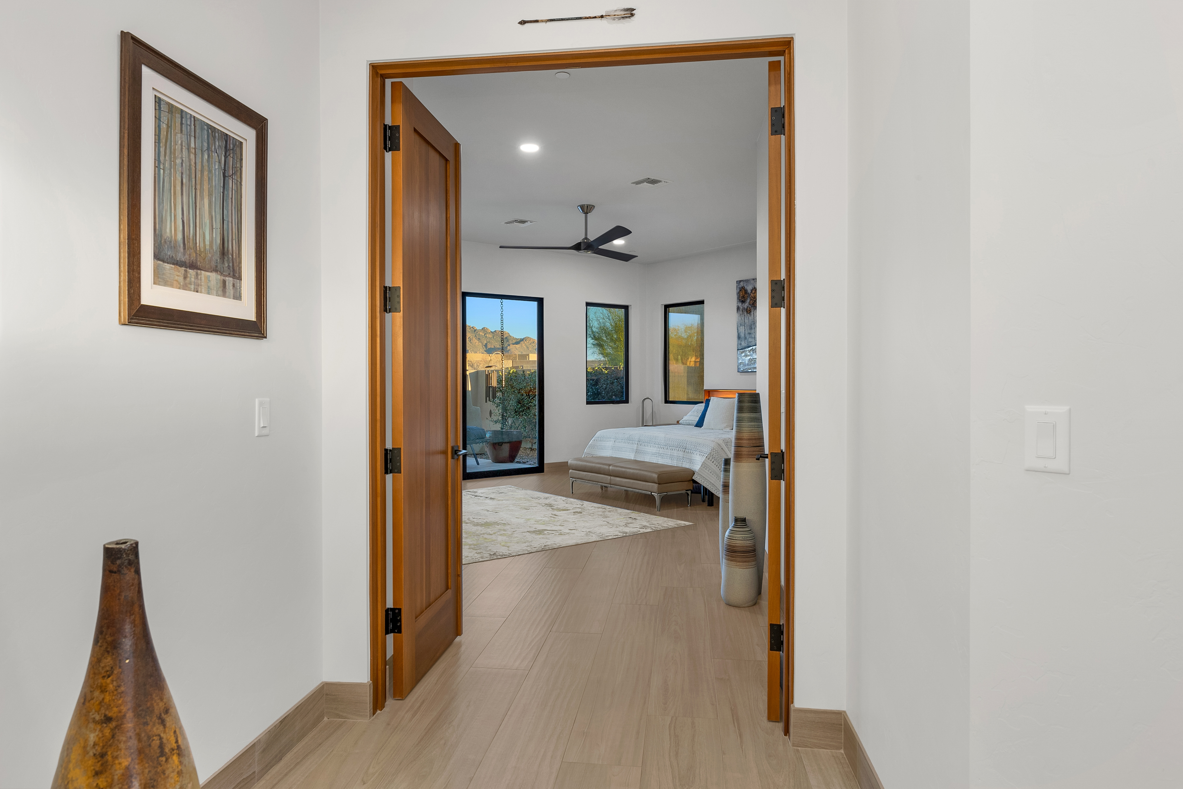 This perspective captures a transition space looking through a set of double wooden doors into a bright, modern bedroom. The foreground features a neutral-toned hallway with light wood-look flooring and a decorative vase, while the bedroom beyond showcases a bed, a bench, and large windows that frame a desert landscape. The clean, minimalist aesthetic and warm wood tones create an inviting and airy atmosphere.