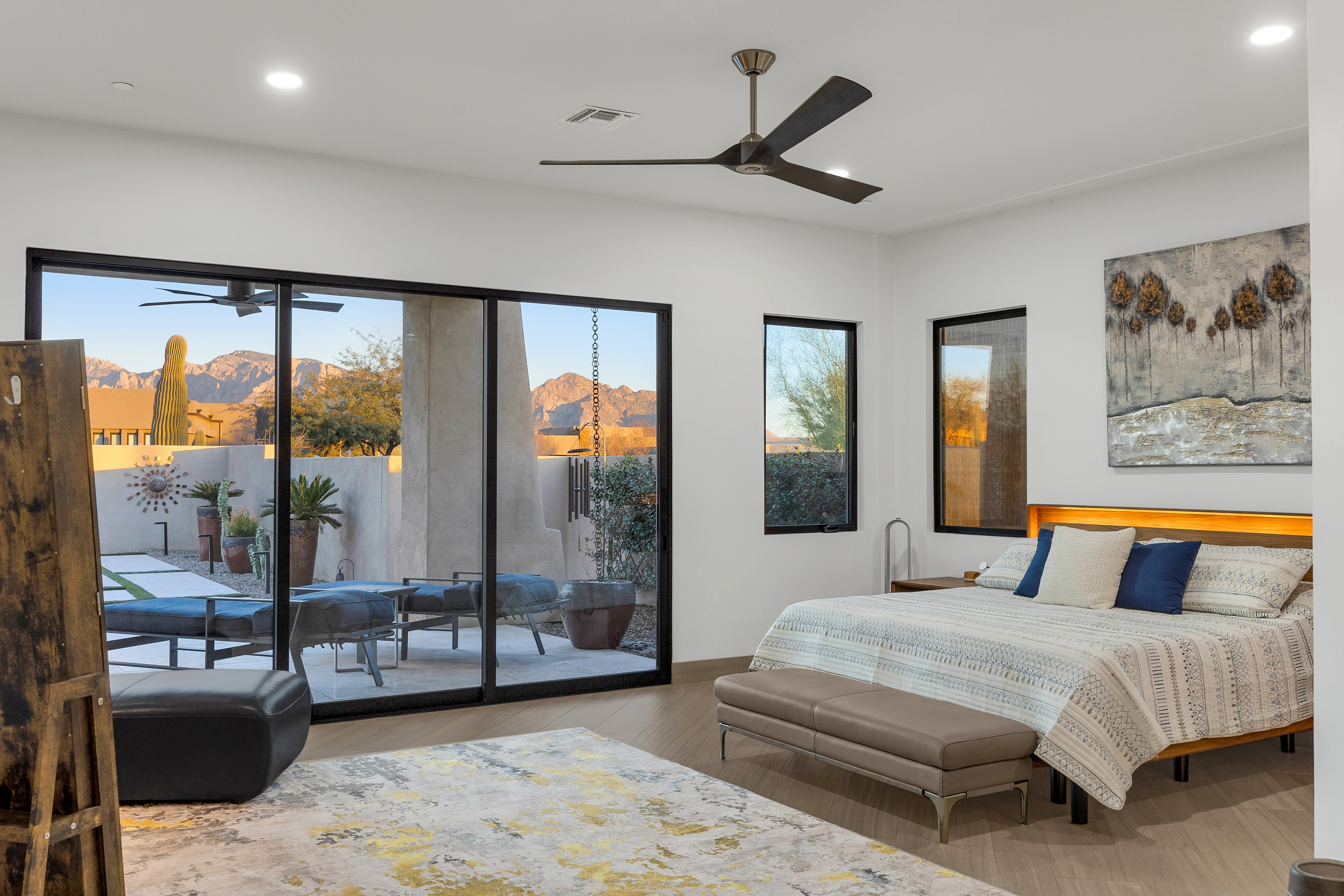 This spacious primary bedroom features a contemporary design with light wood-look flooring and a neutral color palette accented by deep blue pillows. A large sliding glass door provides a seamless transition to a private patio, framing stunning mountain views and desert landscaping. The room is anchored by a bed with an integrated headboard light and a textured piece of wall art, creating a serene and sophisticated atmosphere.