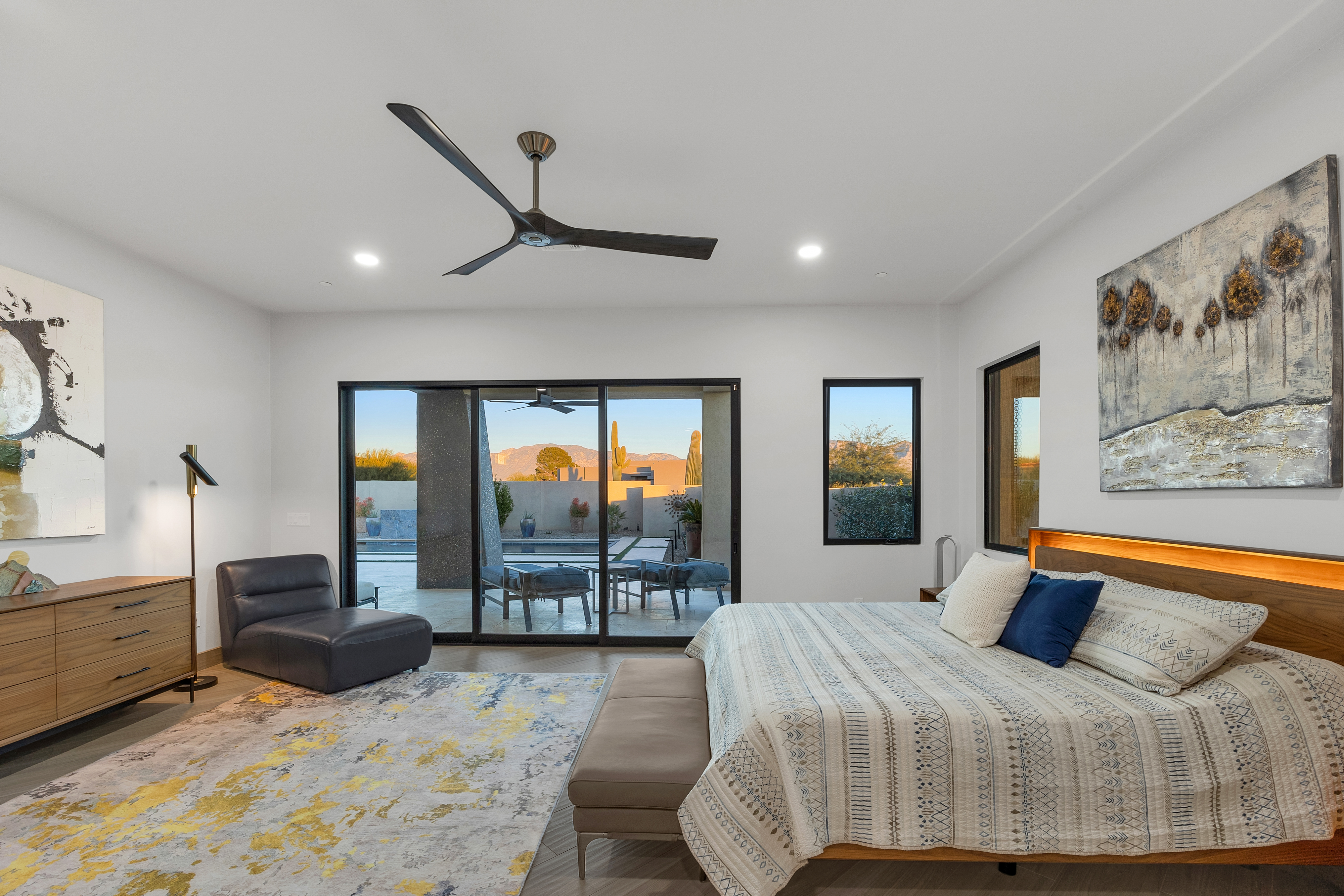 This spacious primary bedroom features a contemporary design with a neutral color palette, accented by a textured area rug and abstract wall art. A large bed with patterned bedding sits against a headboard with integrated lighting, while a sleek leather lounge chair provides a comfortable seating area. Large sliding glass doors offer a seamless transition to the outdoor patio, framing a scenic view of the desert landscape and mountains beyond.