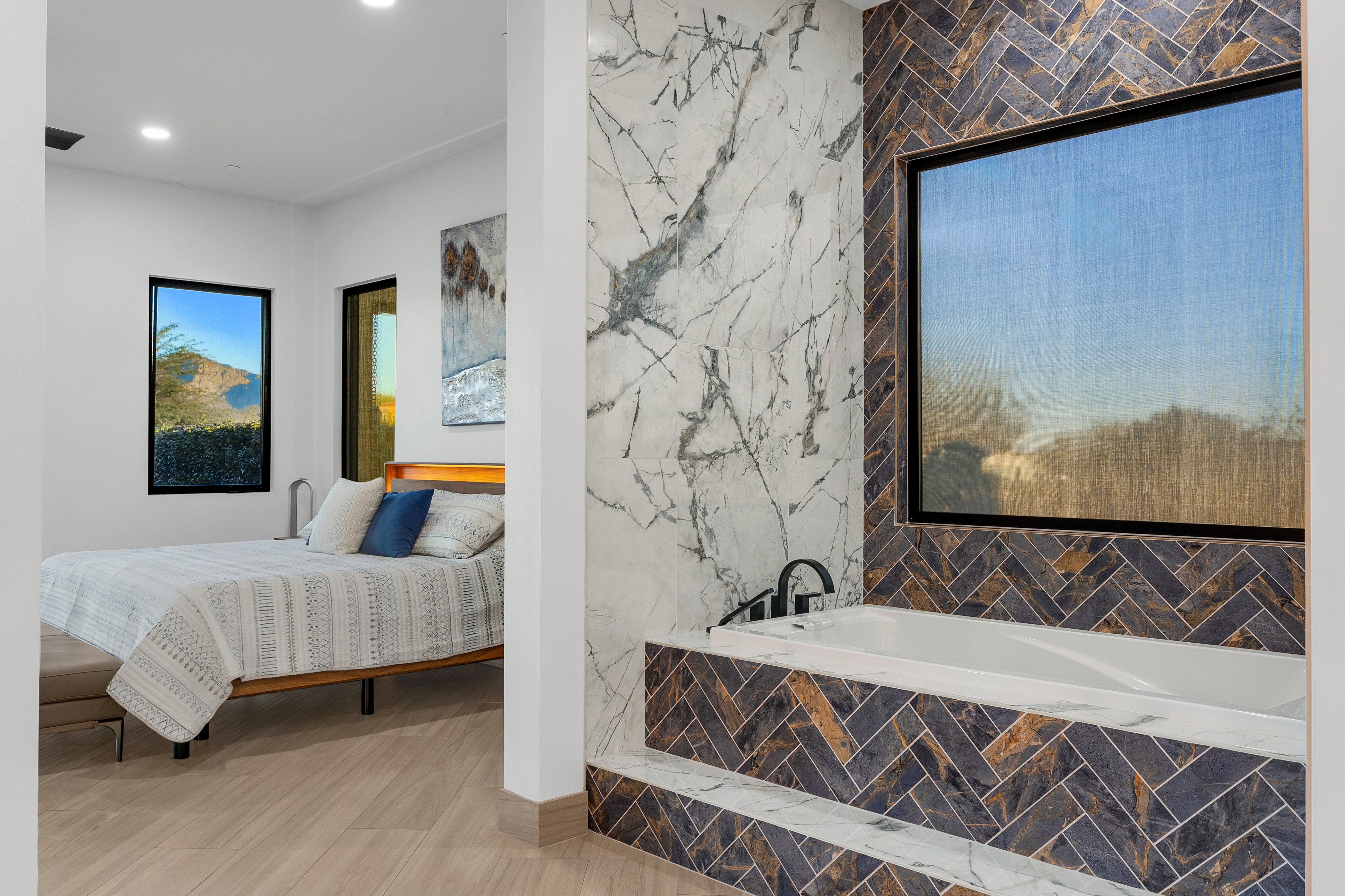 This view captures a primary bathroom and bedroom suite, showcasing a luxurious soaking tub encased in dark, herringbone-patterned tile with a stone-topped step. Adjacent to the tub, a large section of the wall is finished in white, veined stone, creating a striking contrast. Through the open transition, a bedroom is visible, featuring a bed with patterned bedding and a view of the desert landscape through a window.
