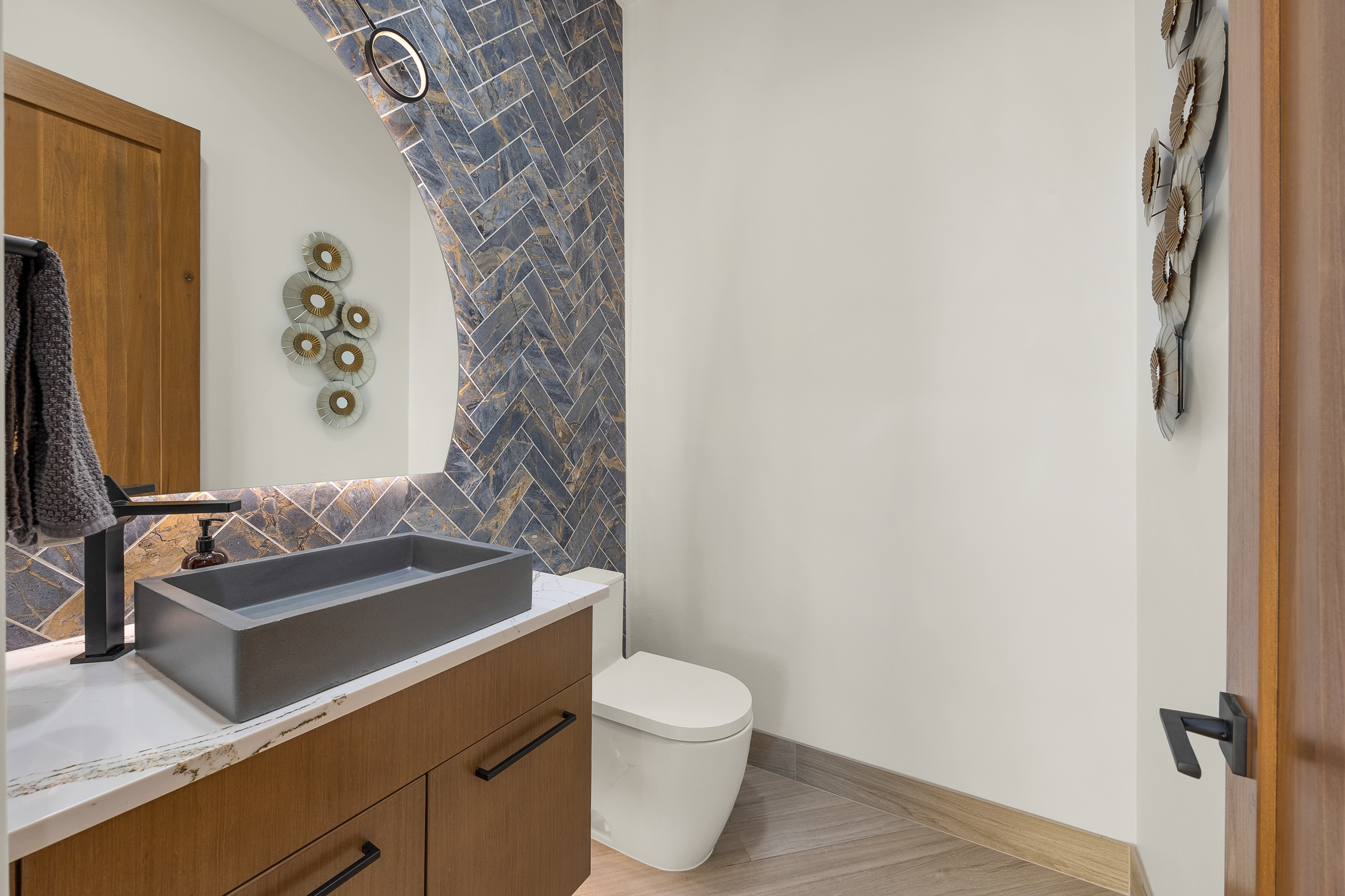 This modern powder room features a striking herringbone-patterned tile accent wall in deep blue and gold tones that frames a large, uniquely shaped mirror. A rectangular, dark-toned vessel sink sits atop a light-colored stone vanity with wood-toned cabinetry, complemented by matte black fixtures. The space is finished with neutral walls and wood-look flooring, creating a sophisticated and contemporary aesthetic.