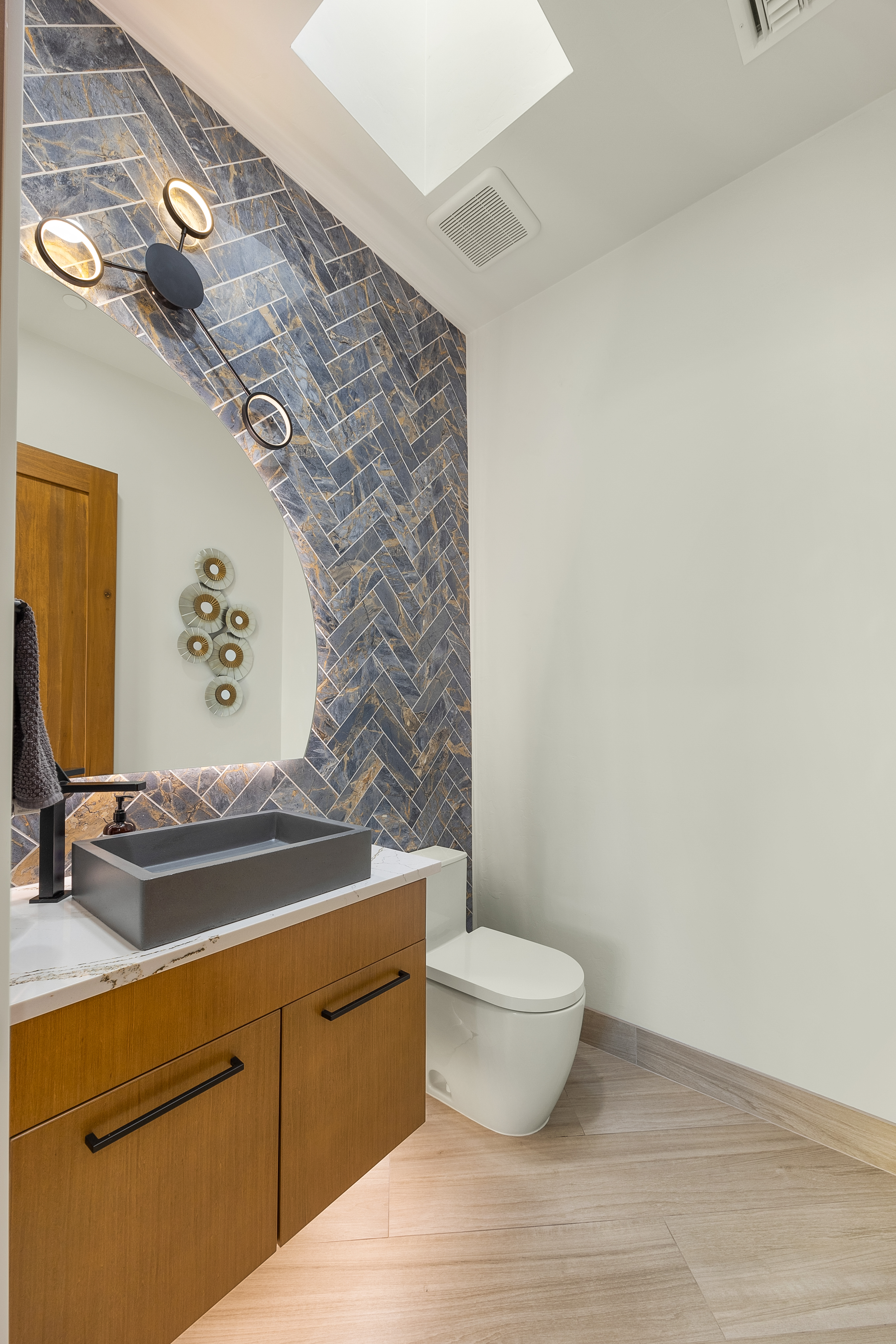 This modern powder room features a striking accent wall with blue and gold patterned tile laid in a herringbone pattern. A floating wood vanity with a dark rectangular vessel sink and matte black hardware sits beneath a uniquely shaped mirror, while a white toilet is positioned to the side. The space is illuminated by a contemporary multi-arm light fixture and a skylight, creating a bright and sophisticated atmosphere.