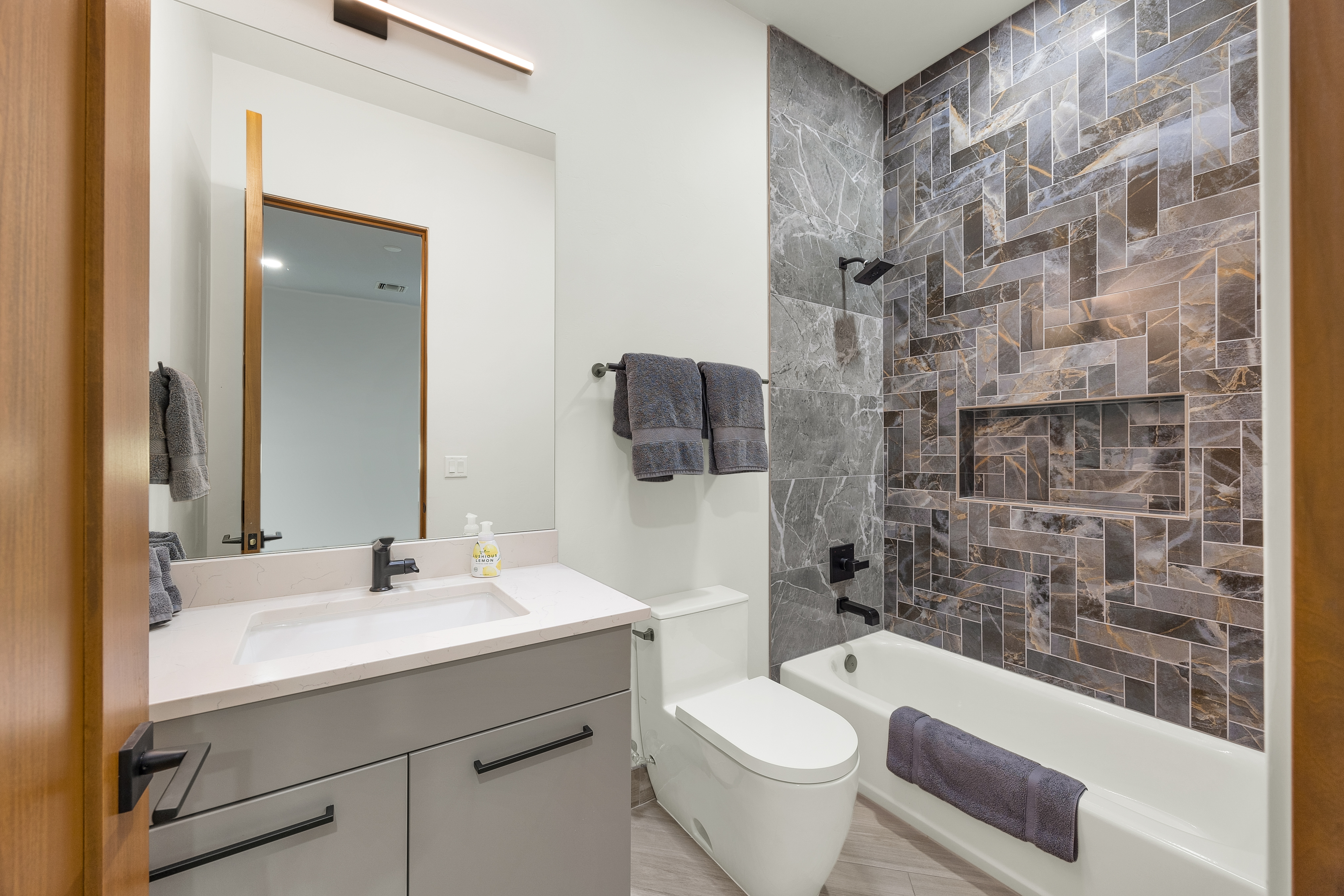 This modern bathroom features a sleek gray vanity with a white countertop and a rectangular undermount sink, complemented by matte black hardware. The shower area is a standout feature, showcasing a striking herringbone-patterned tile surround in dark, multi-tonal stone with a built-in recessed niche. A white toilet and a large, frameless mirror with a minimalist light fixture complete the clean, contemporary aesthetic of the space.