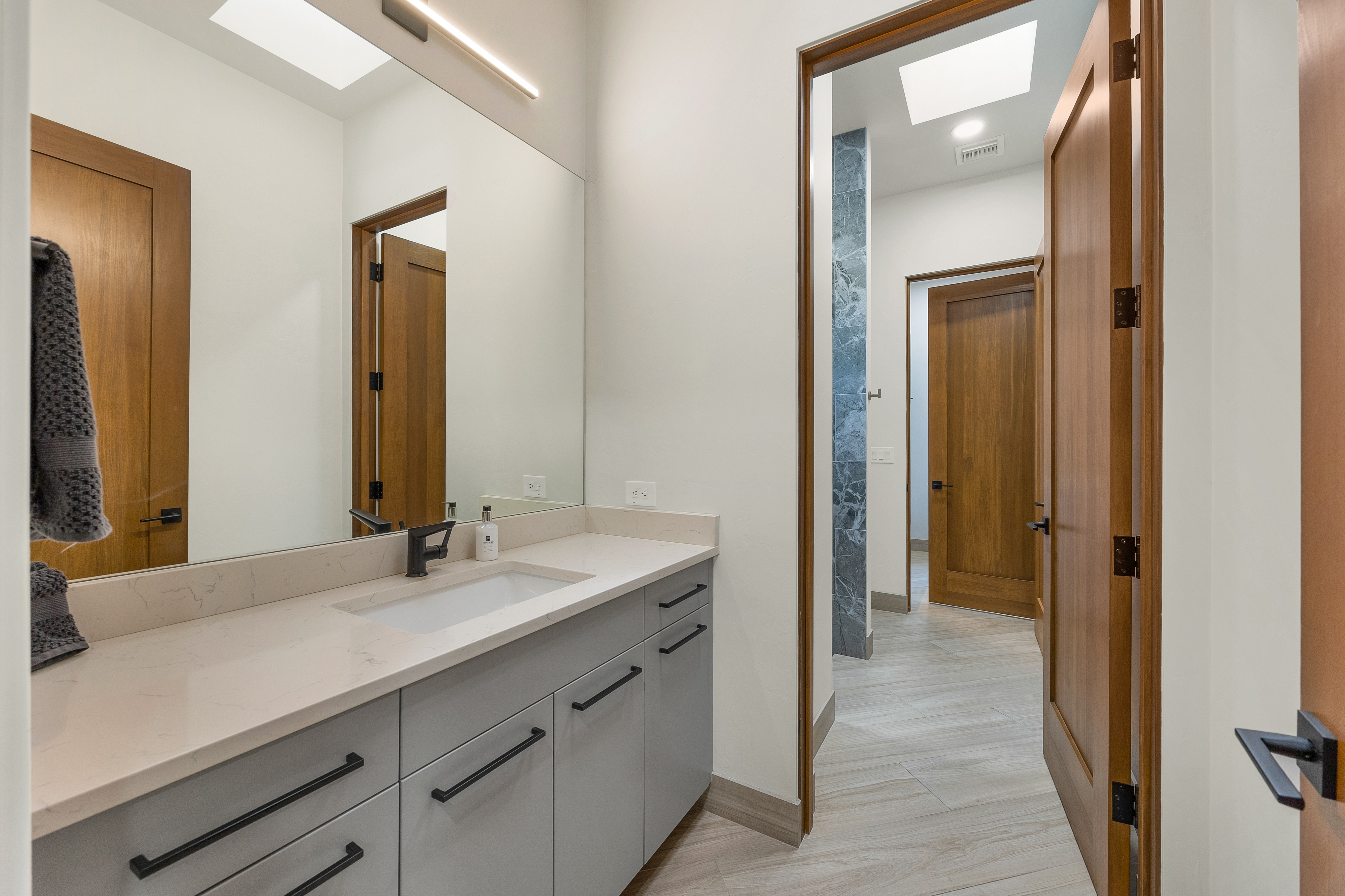 This modern bathroom features a sleek vanity with light gray cabinetry, a stone countertop, and a matte black faucet. A large, frameless mirror reflects the hallway and an adjacent doorway, while the space is illuminated by a minimalist light fixture and a skylight. The neutral color palette and clean lines create a contemporary and functional aesthetic.