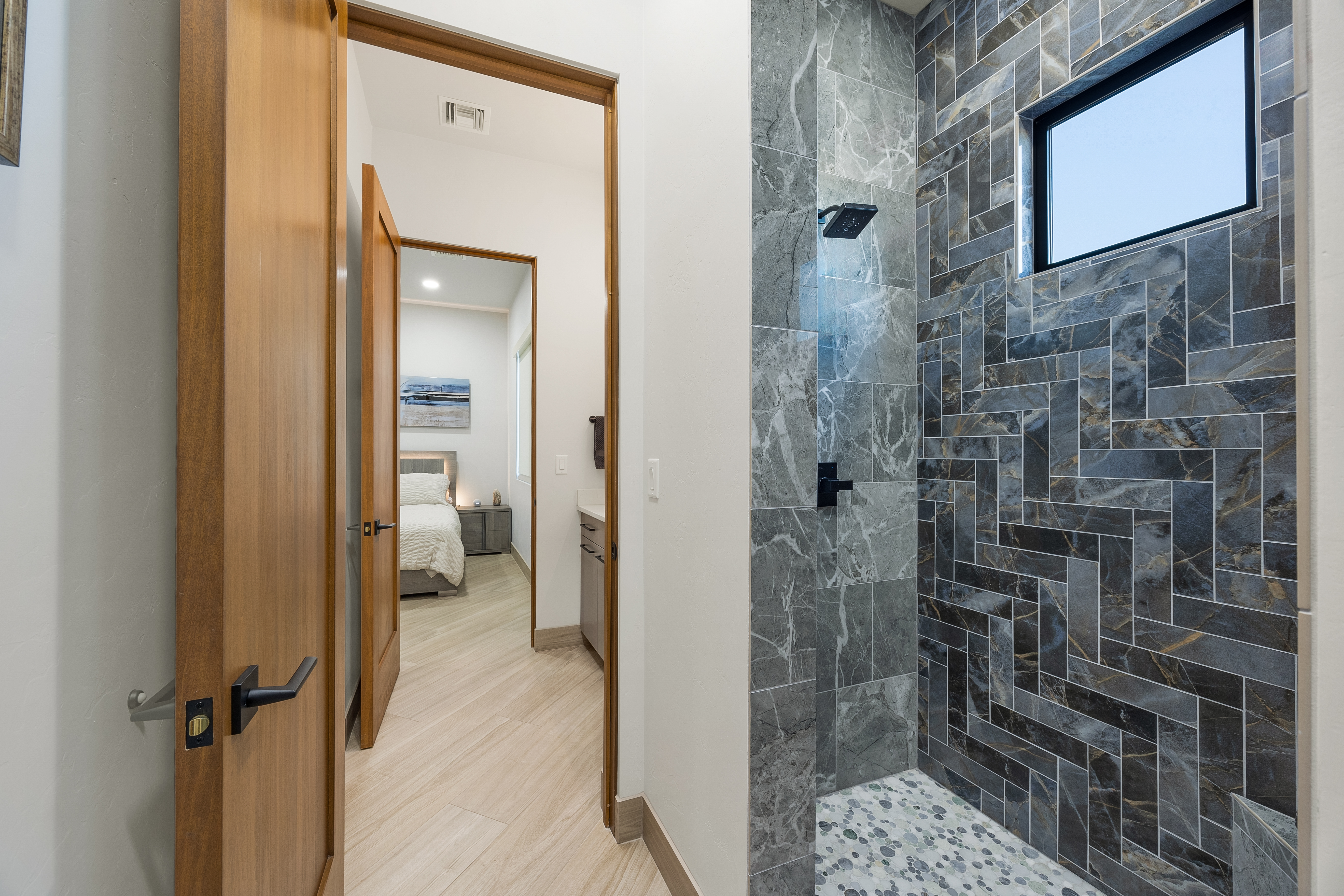 This interior view captures a modern bathroom featuring a walk-in shower with a striking herringbone-patterned dark stone tile wall and a pebble-style floor. The perspective looks through a wooden doorway into an adjacent bedroom, creating a sense of flow between the spaces. The neutral color palette and clean lines contribute to a contemporary and sophisticated aesthetic.