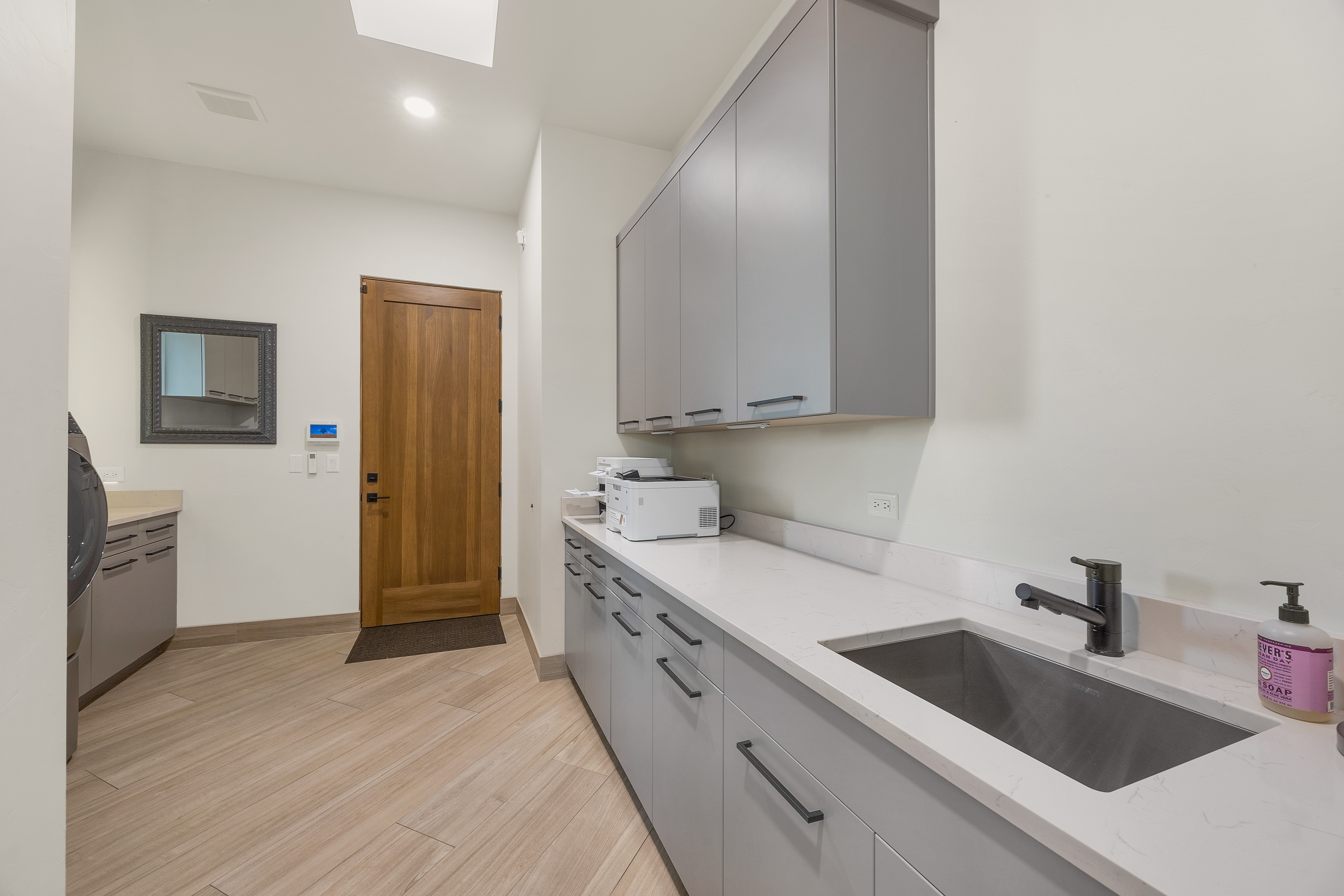 This functional laundry room features sleek gray cabinetry, a white countertop with an integrated sink, and a modern black faucet. The space includes a wood-toned door, light-colored wood-look flooring, and a printer placed on the counter, suggesting a multi-purpose utility area. The perspective is from the doorway, looking across the room toward the cabinetry and the adjacent laundry appliances.