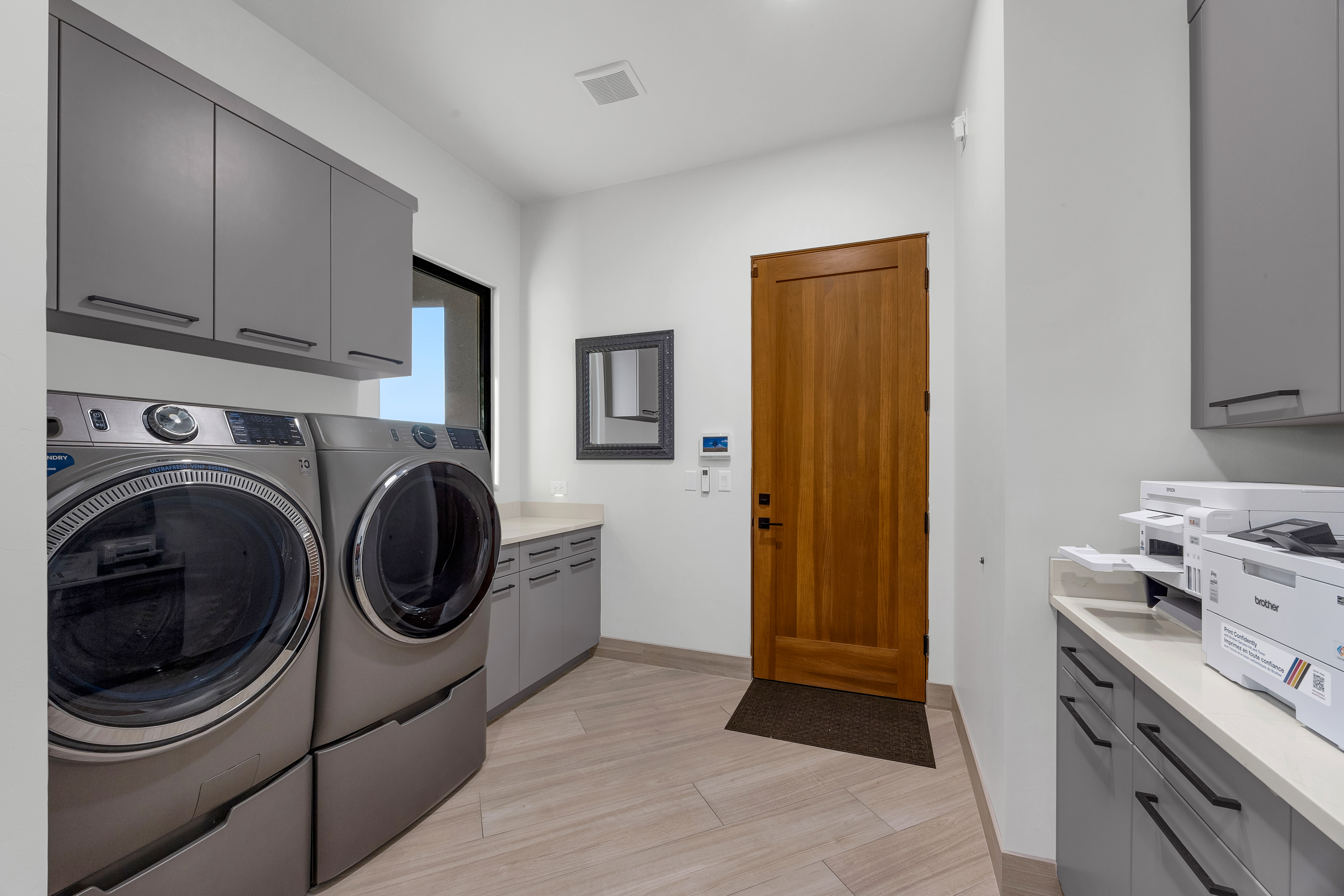 This functional laundry room features a modern aesthetic with gray cabinetry, light-toned wood-look flooring, and a prominent wooden door. The space is equipped with a side-by-side washer and dryer set, a built-in countertop for folding, and a printer station on the opposite side. A small mirror and a wall-mounted control panel add utility to the clean, well-lit room.