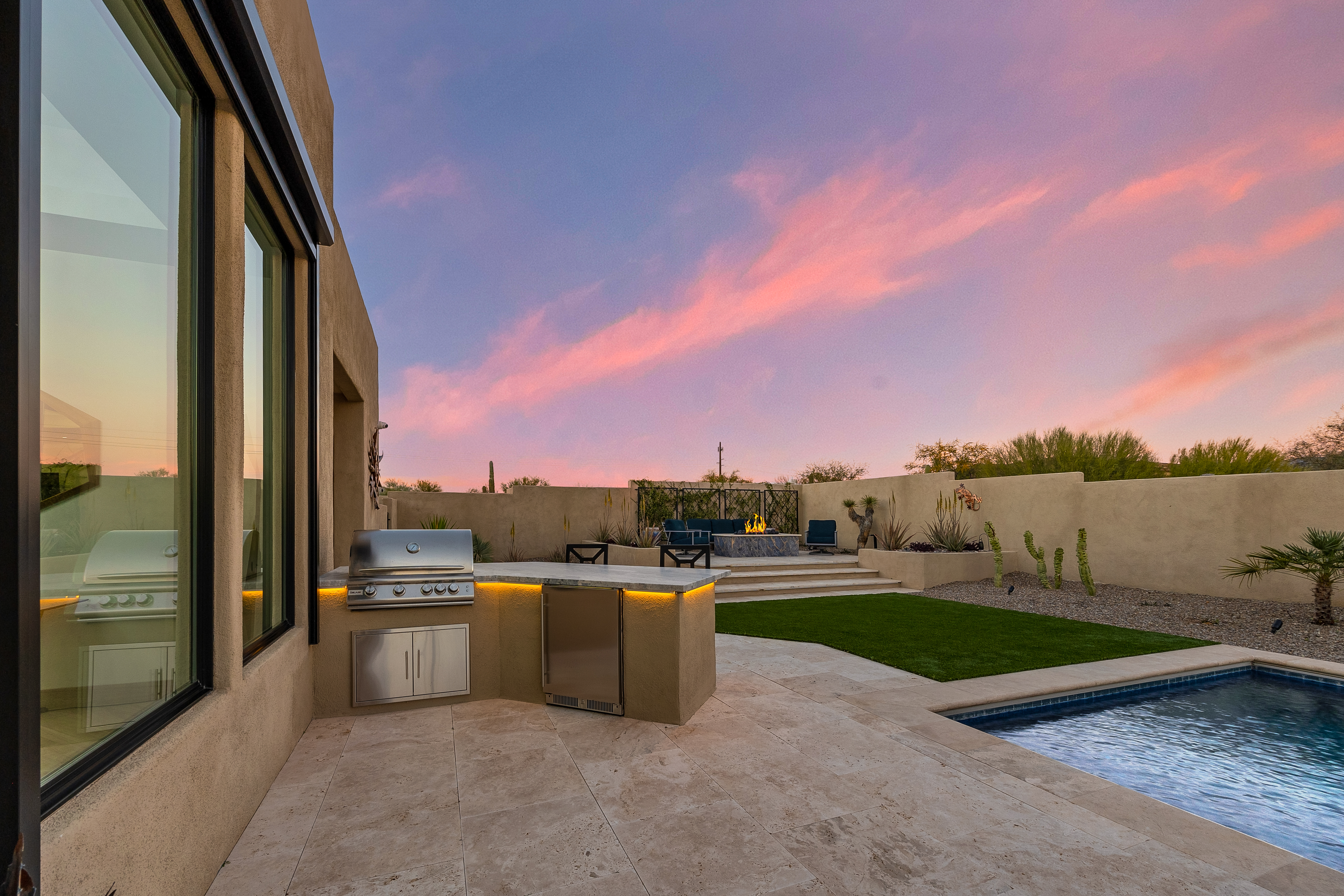 This outdoor living space features a built-in kitchen area with a stainless steel grill and under-counter refrigerator, illuminated by warm LED accent lighting. The patio transitions into a manicured lawn and a swimming pool, with a raised fire pit area set against a desert-style wall. The scene is captured at sunset, with a vibrant pink and blue sky providing a serene backdrop to the modern, low-maintenance landscape.