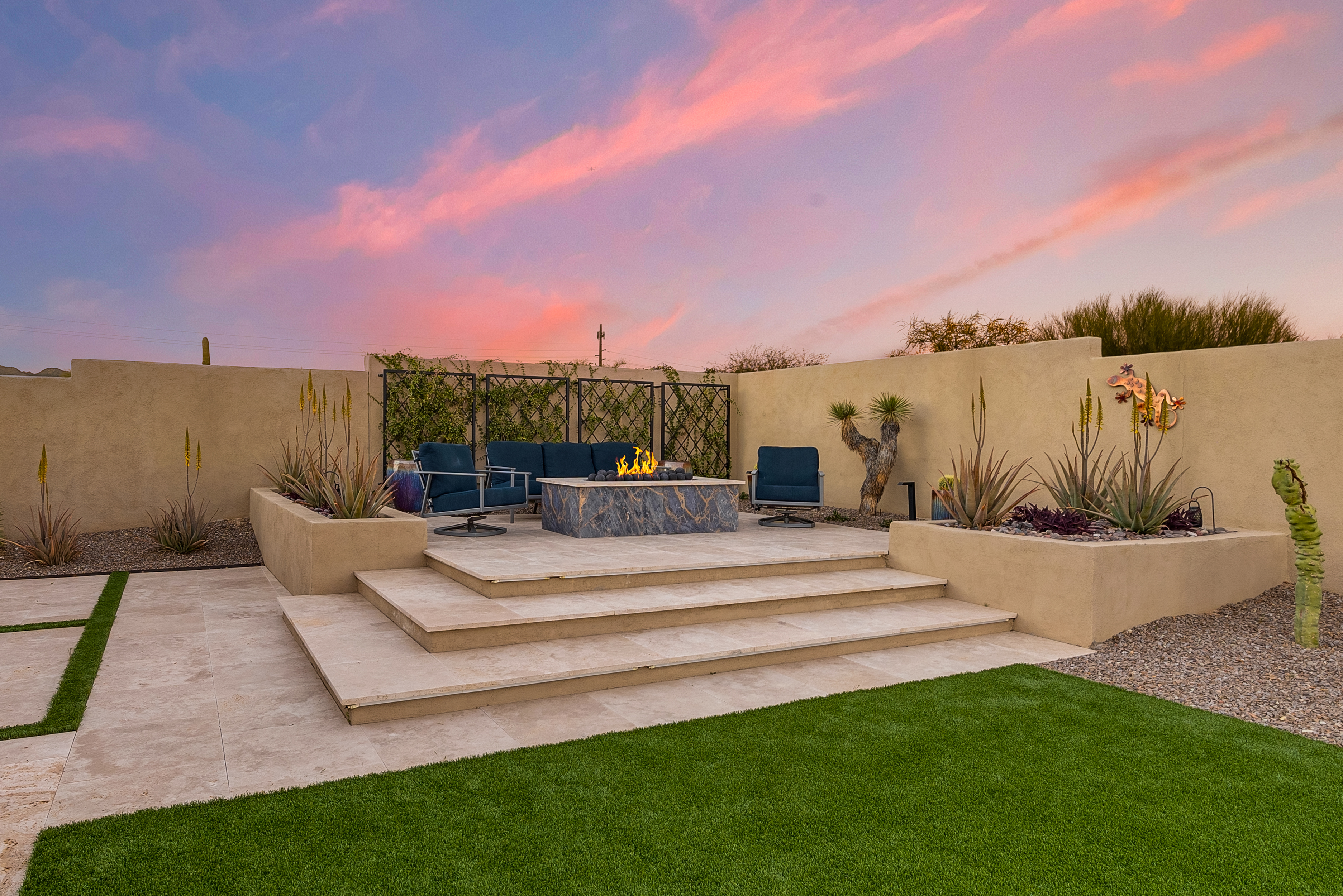This inviting outdoor living space features a raised stone patio with a central fire pit and comfortable lounge seating. The area is framed by desert-inspired landscaping, including built-in planters and a decorative wall, all set against a vibrant sunset sky. The combination of clean stone surfaces and artificial turf creates a low-maintenance, modern aesthetic perfect for evening relaxation.