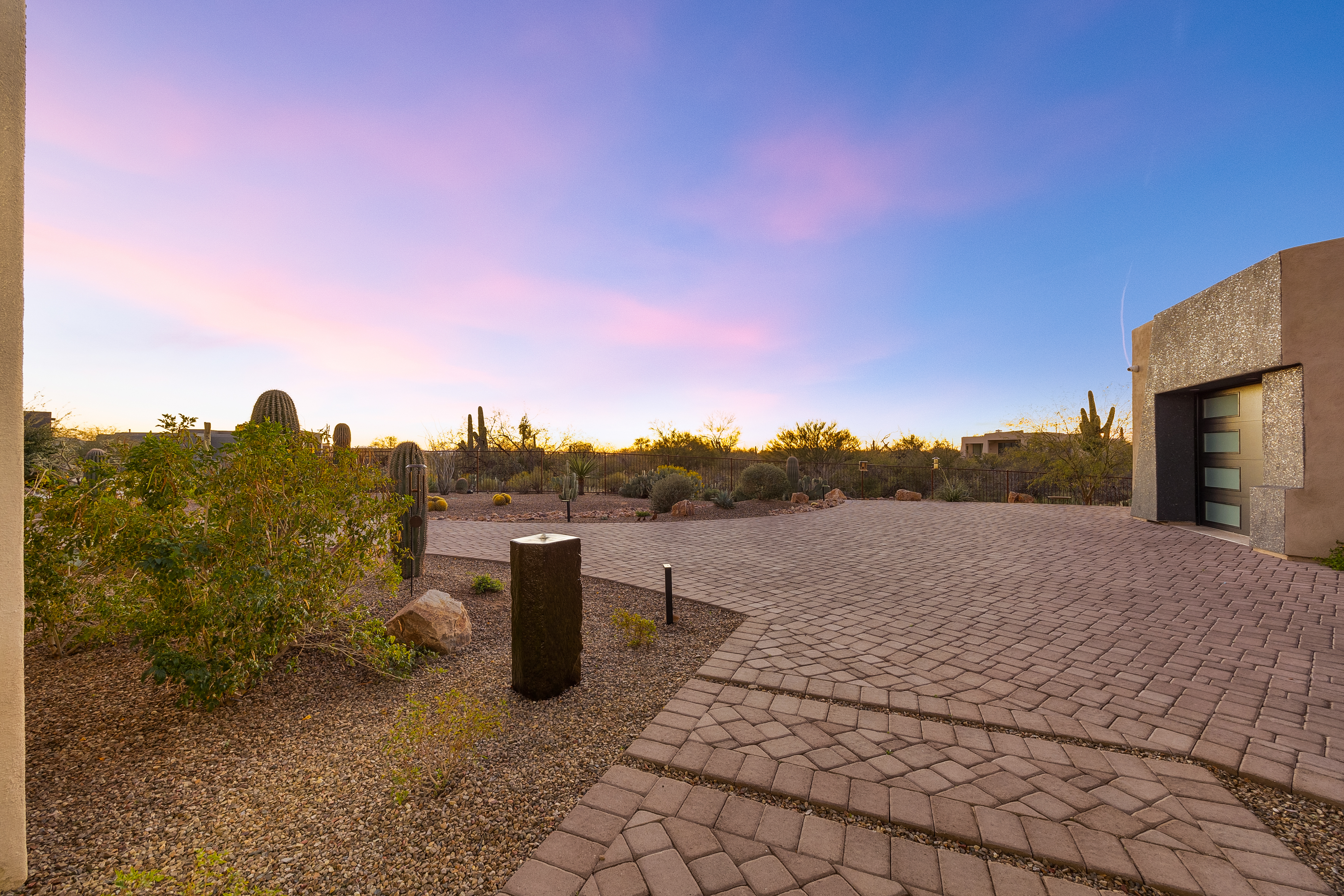 This desert-style entryway features a wide, paved driveway leading to a modern home entrance with a sleek, multi-panel door. The landscape is characterized by native cacti, gravel ground cover, and a minimalist water feature, all set against a vibrant sunset sky. The perspective captures the expansive, open feel of the property, emphasizing its integration with the surrounding arid environment.