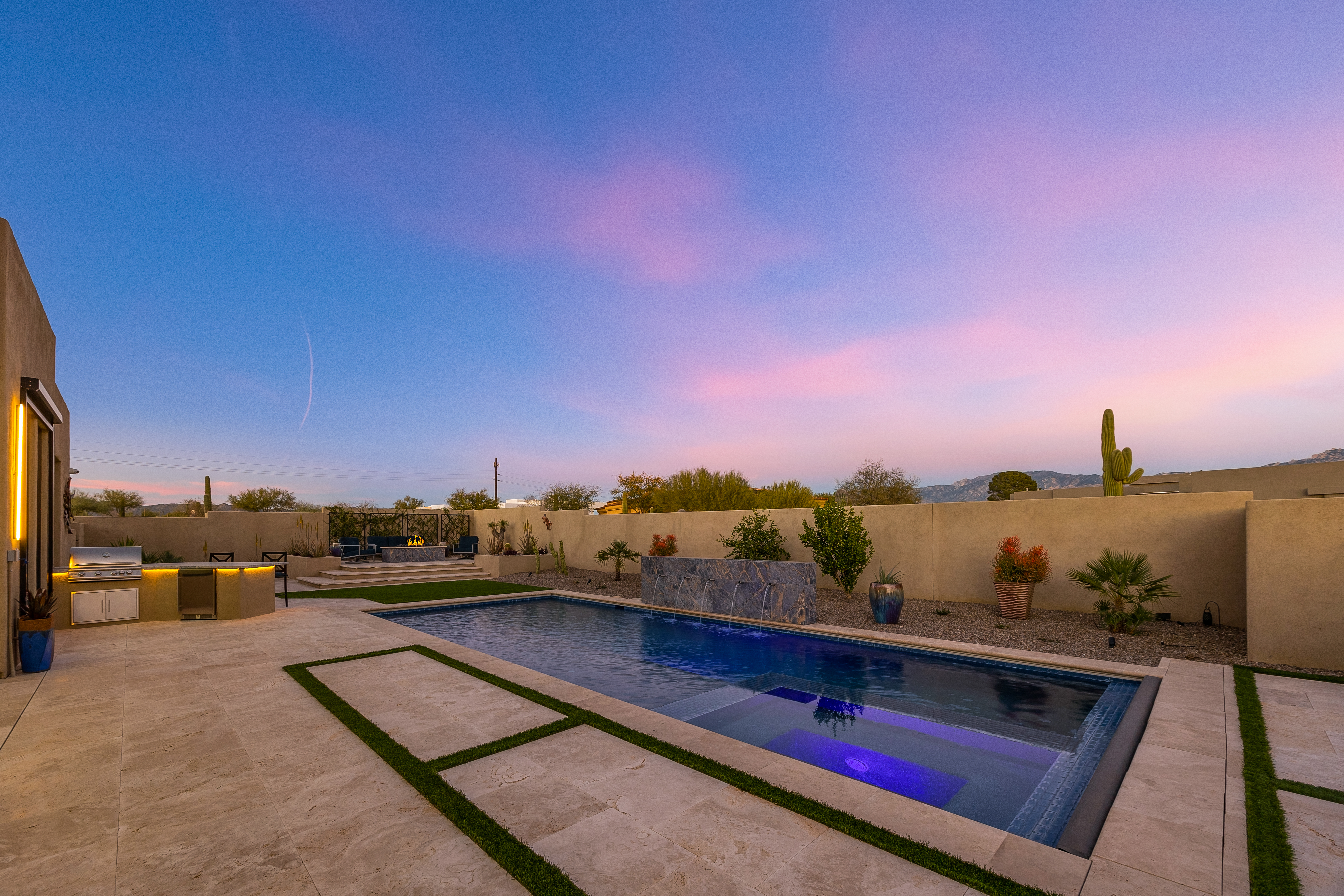This stunning desert backyard features a sleek, rectangular swimming pool with an integrated spa and a modern water feature wall. The surrounding patio is composed of large stone pavers with integrated turf strips, complemented by an outdoor kitchen area and a raised fire pit lounge. The scene is captured at dusk, with the vibrant pink and blue sunset sky casting a serene glow over the contemporary landscape.