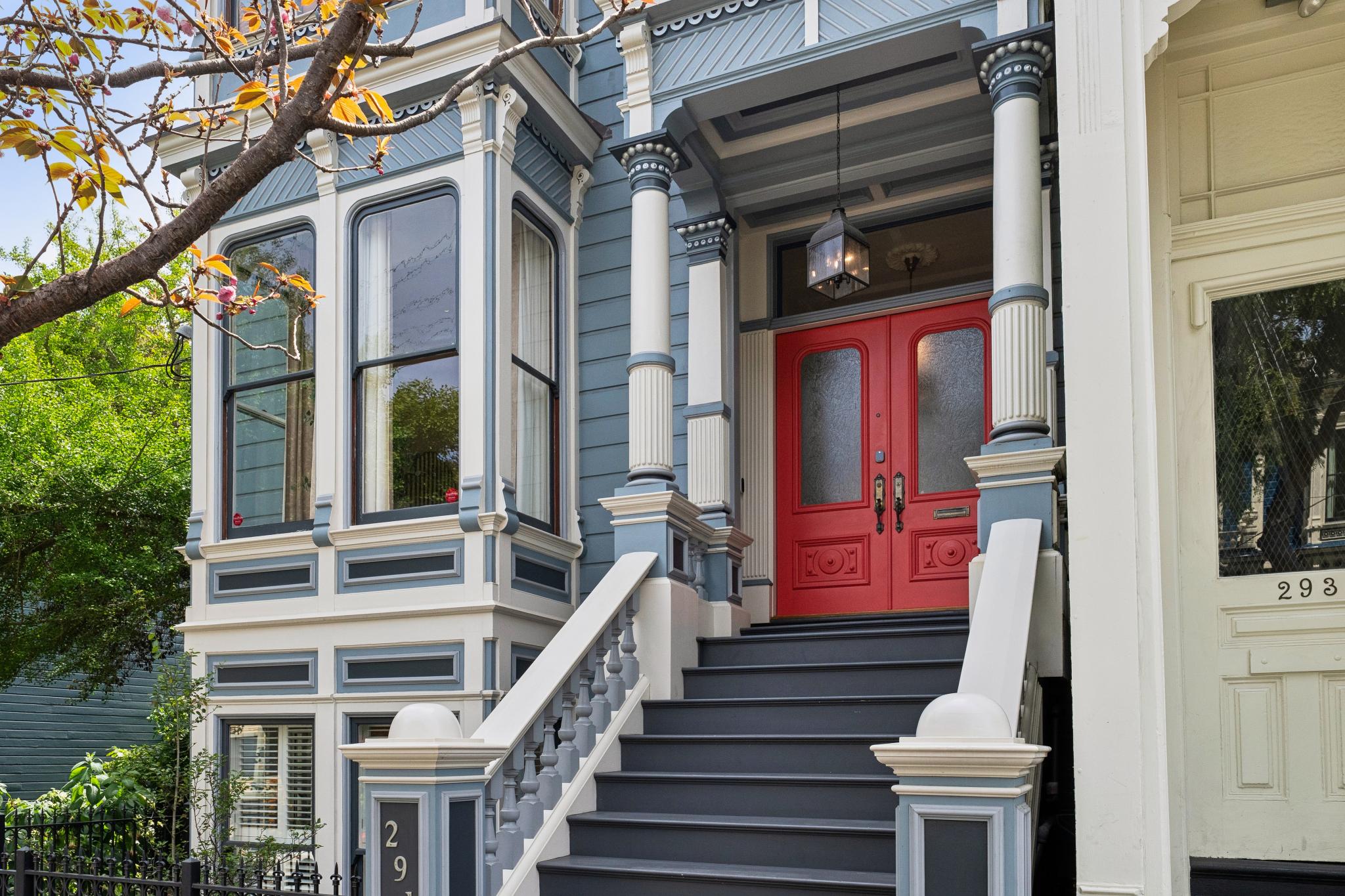 This charming Victorian-style home features a striking red double door entryway framed by ornate white columns and blue-grey siding. The perspective captures the inviting front staircase leading up to the porch, highlighting the intricate architectural details and classic San Francisco aesthetic. The scene is bright and well-maintained, emphasizing the property's curb appeal and historic character.