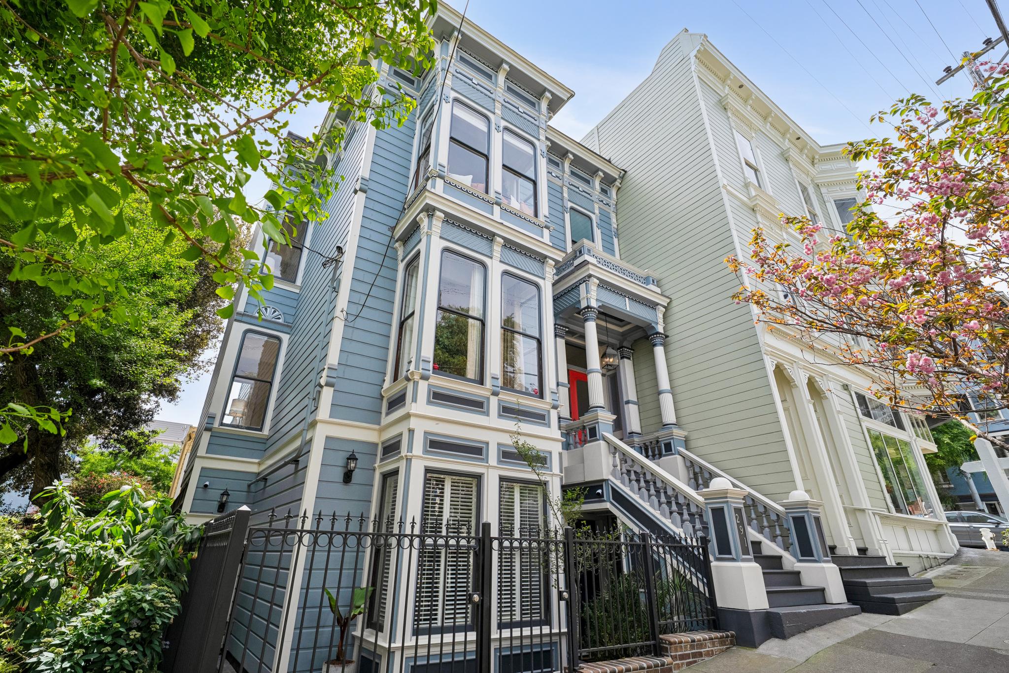 This striking Victorian-style home features a classic blue facade with intricate white trim and a prominent bay window structure. The property is elevated with a grand staircase leading to a vibrant red front door, framed by lush greenery and a blooming cherry blossom tree. The low-angle perspective emphasizes the architectural height and historical charm of this quintessential San Francisco-style residence.