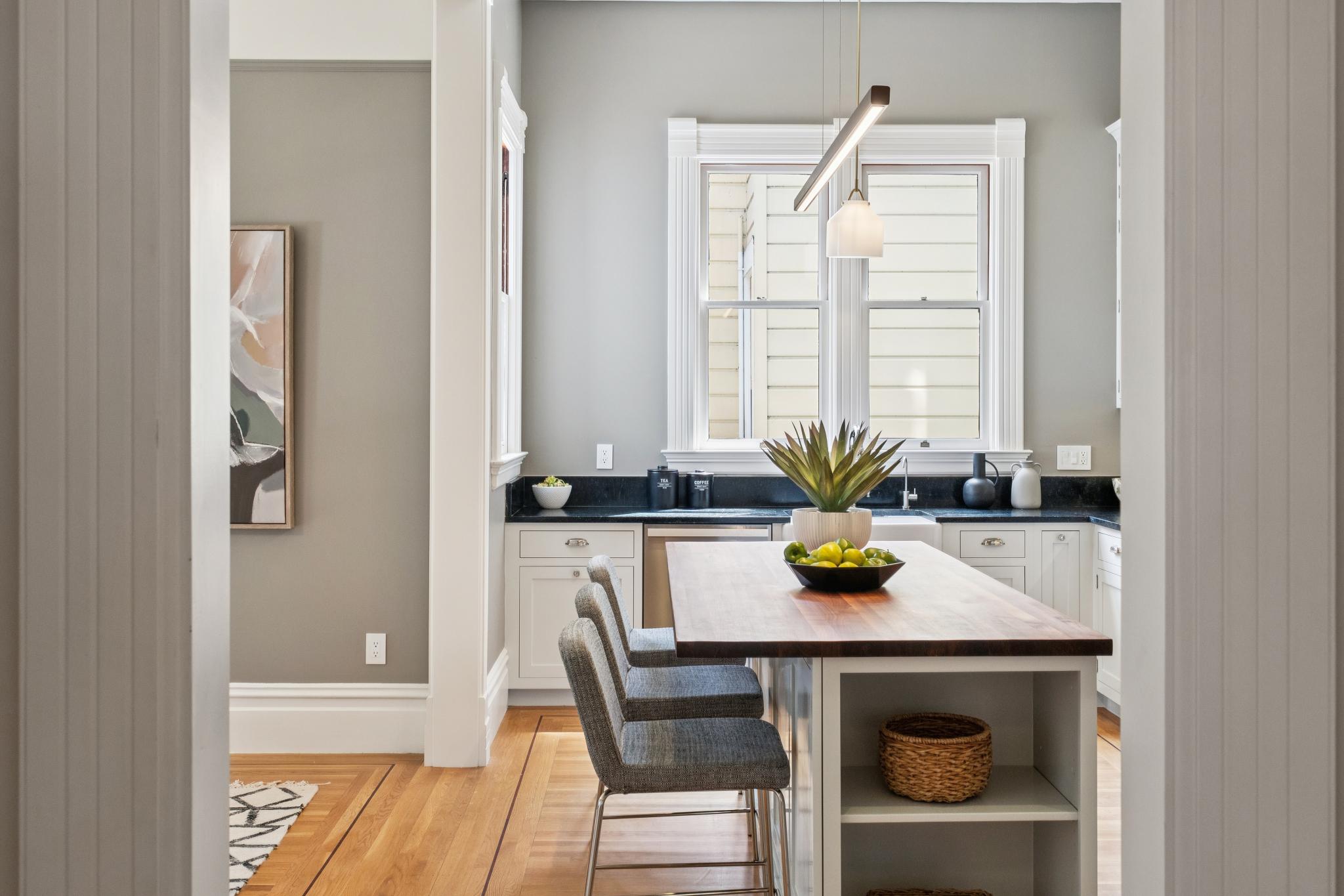 This bright and inviting kitchen features a central island with a rich wood countertop and gray base cabinetry, paired with three modern gray upholstered bar stools. The space is accented by white cabinetry, dark stone countertops, and a large window that brings in natural light, all set against soft gray walls and warm hardwood flooring. The perspective is framed through a doorway, creating a cinematic depth that highlights the clean, transitional design of the home.