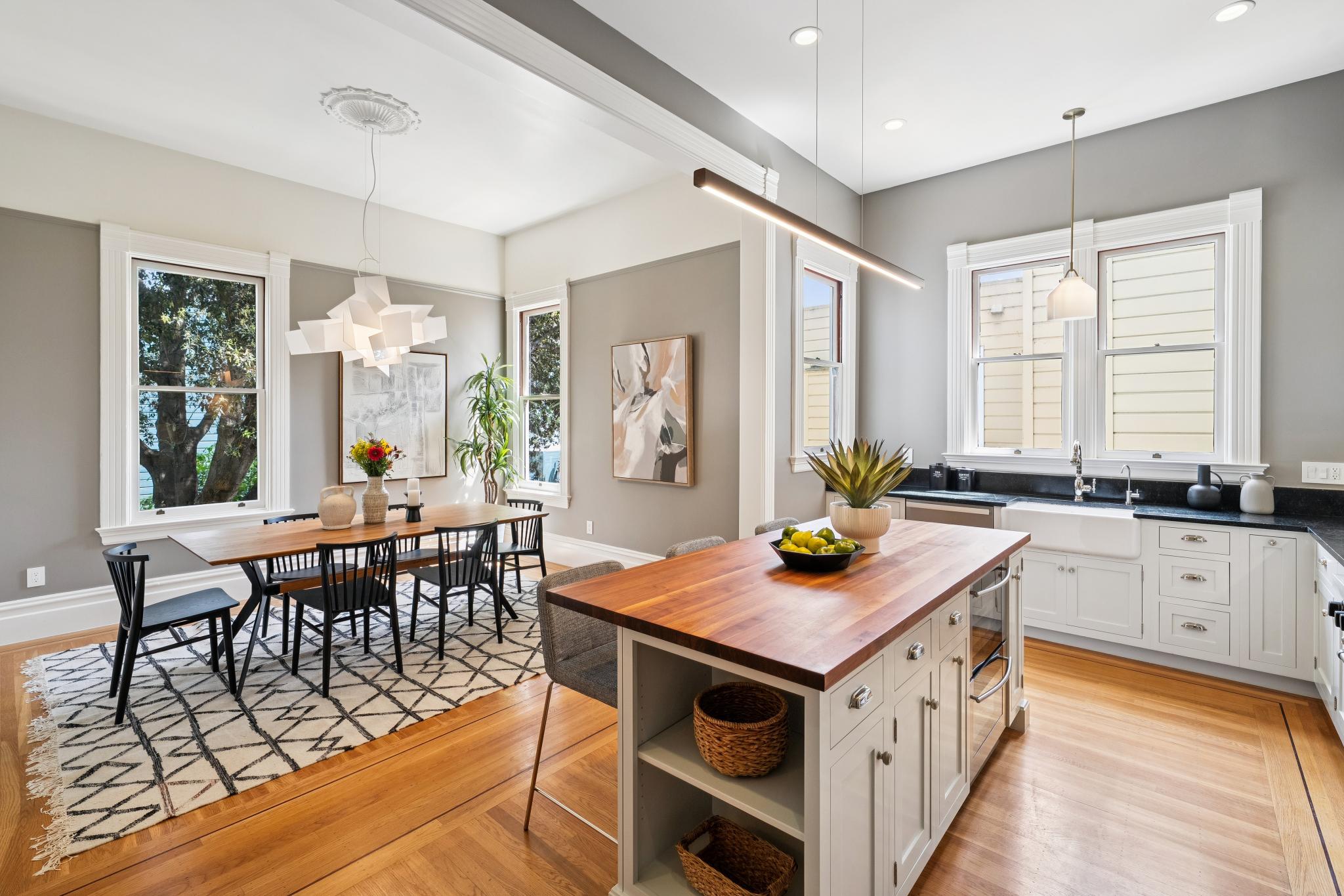 This bright and airy open-concept kitchen and dining area features a large butcher-block island, white cabinetry, and a farmhouse sink. The dining space is anchored by a modern geometric chandelier and a patterned area rug, creating a sophisticated yet inviting atmosphere. The warm hardwood floors and neutral grey walls provide a cohesive, high-end aesthetic perfect for modern living.