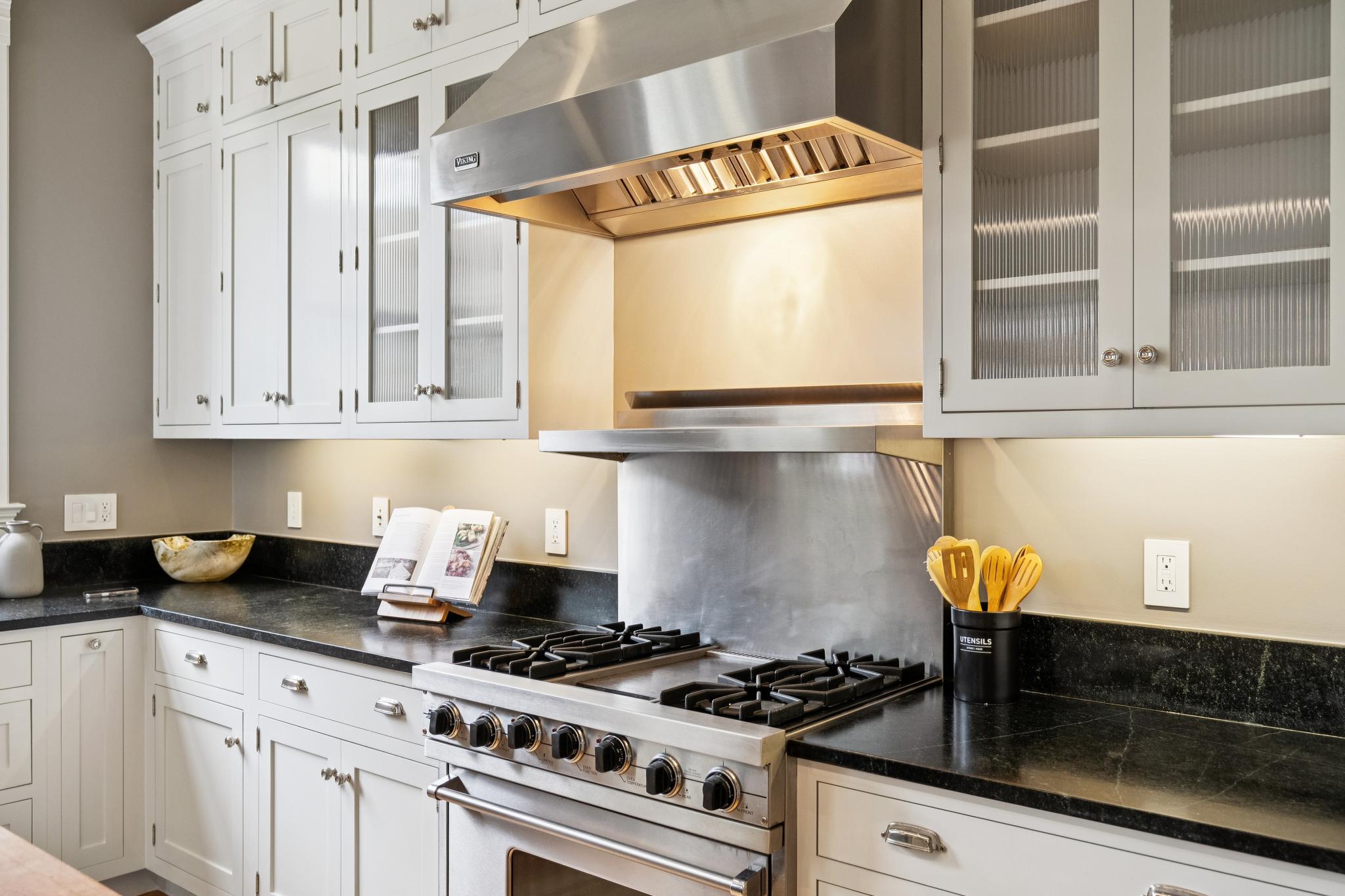 This elegant kitchen features classic white cabinetry with glass-fronted upper cabinets and a striking black granite countertop. A professional-grade stainless steel Viking range and matching range hood serve as the focal point, complemented by a sleek stainless steel backsplash. The space is well-lit with under-cabinet lighting, creating a warm and inviting atmosphere perfect for culinary enthusiasts.