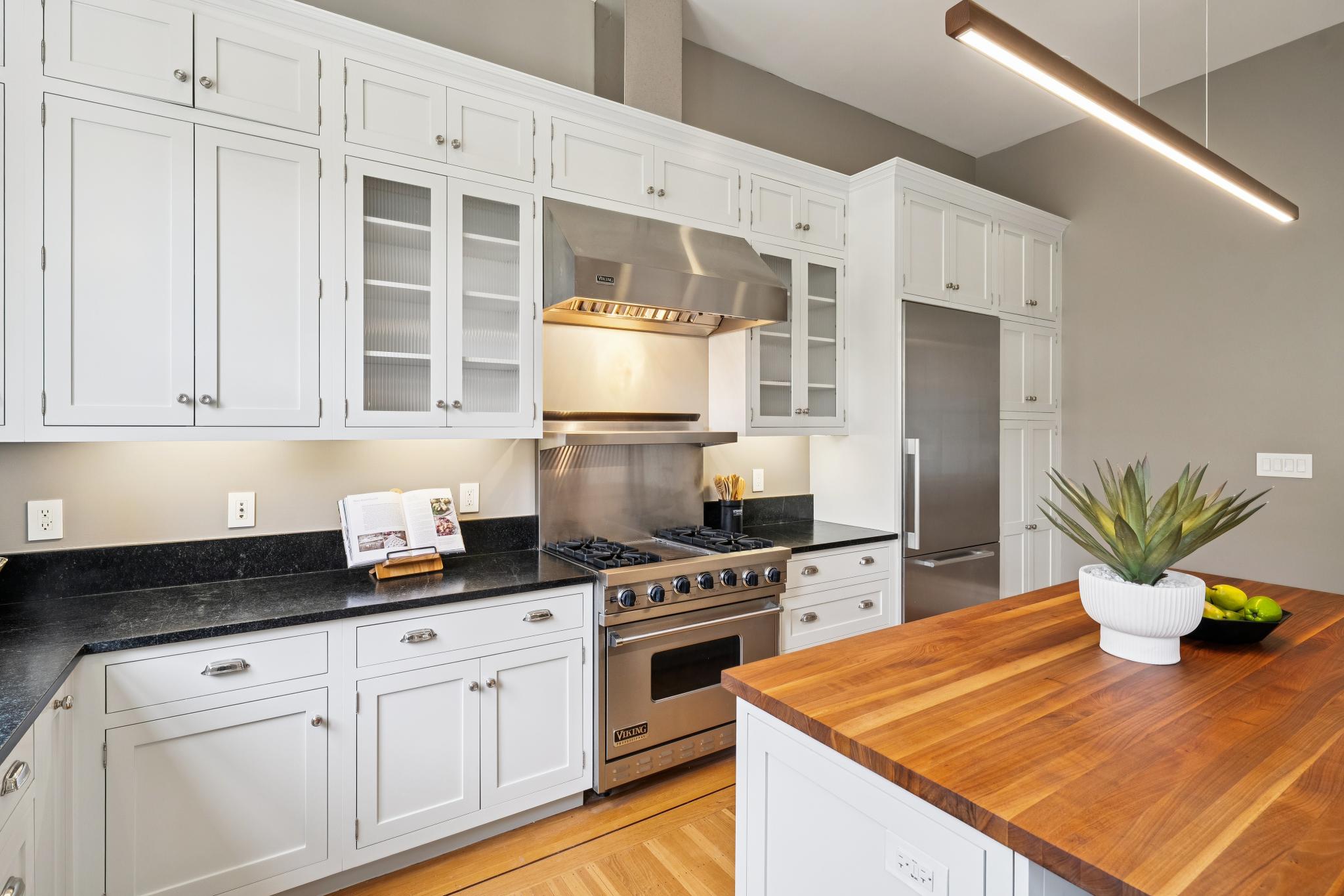 This bright and elegant kitchen features crisp white cabinetry paired with contrasting black granite countertops and a warm wood-topped island. A professional-grade stainless steel Viking range and hood serve as the focal point, complemented by a modern linear pendant light fixture. The space is well-lit and organized, offering a sophisticated blend of classic design and functional culinary workspace.