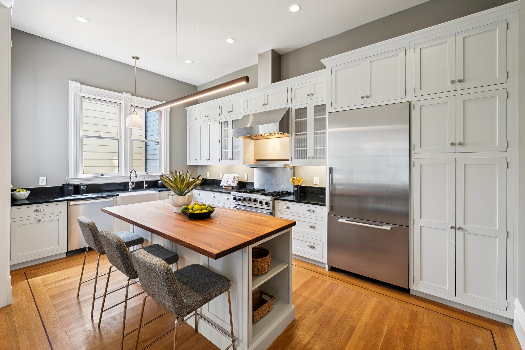 This bright and elegant kitchen features classic white cabinetry, a contrasting dark countertop, and a warm wood-topped center island with bar seating. The space is illuminated by a sleek, modern linear pendant light and recessed lighting, creating a welcoming atmosphere. The stainless steel appliances and hardwood flooring add a touch of sophistication to this well-appointed, functional culinary space.