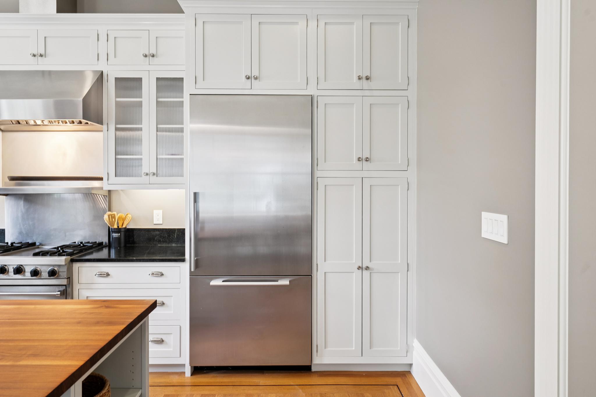 This bright and clean kitchen features classic white shaker-style cabinetry paired with sleek stainless steel appliances, including a large refrigerator and a professional-grade range. A dark granite countertop provides a sharp contrast to the white cabinets, while a warm wood-topped island adds a touch of natural texture to the space. The perspective is a straight-on eye-level shot, highlighting the functional layout and high-end finishes of this modern culinary area.