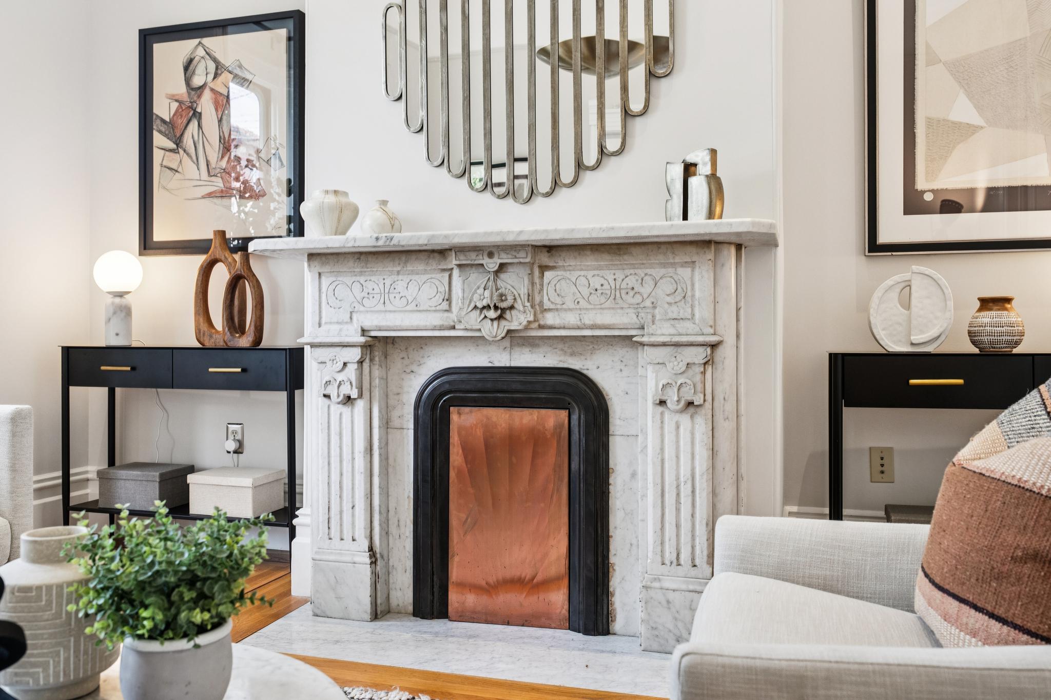 This elegant living room features a stunning, ornate marble fireplace as its central focal point, accented by a unique copper firebox insert. The space is styled with a sophisticated blend of modern and classic elements, including a sculptural mirror above the mantel, sleek black console tables, and plush neutral-toned seating. The composition offers a warm, inviting atmosphere with a refined aesthetic suitable for high-end residential listings.