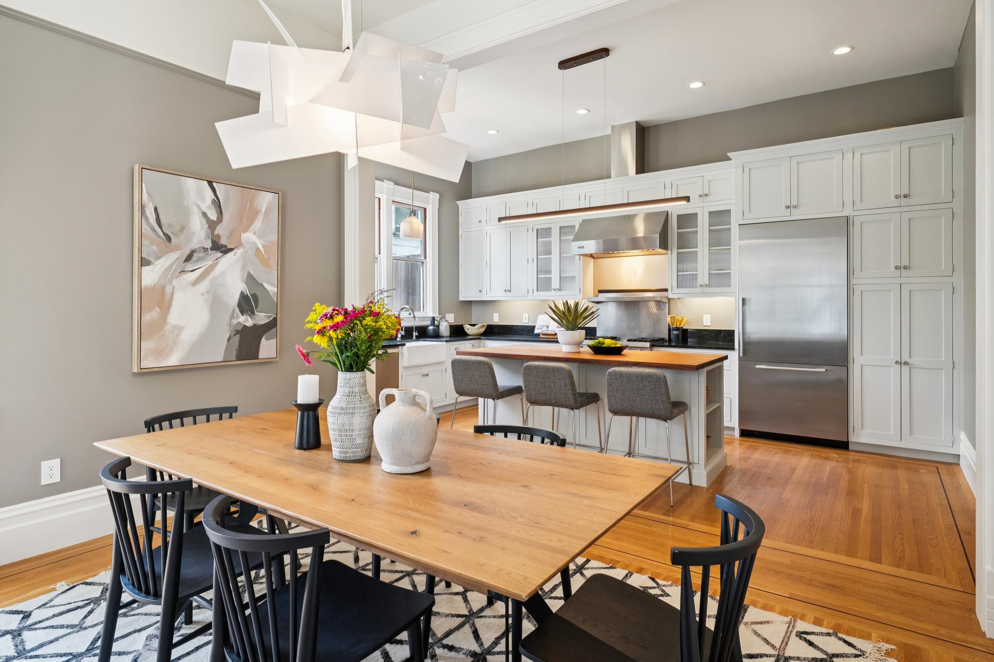 This bright and open-concept space seamlessly blends a dining area with a modern kitchen, featuring a large wooden dining table and a matching kitchen island with bar seating. The kitchen is equipped with white cabinetry, stainless steel appliances, and a sleek, dark countertop, all illuminated by contemporary pendant lighting. The warm hardwood floors and neutral wall tones create an inviting, sophisticated atmosphere perfect for both daily living and entertaining.