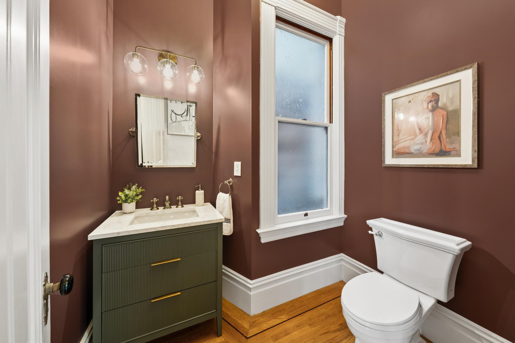 This elegant powder room features rich, deep brown walls that contrast beautifully with a sage green vanity and white marble countertop. The space is accented by gold-toned hardware, a modern three-light fixture, and a framed piece of art, creating a sophisticated and moody atmosphere. The room is completed by a white toilet and classic white trim, offering a polished and high-end aesthetic.