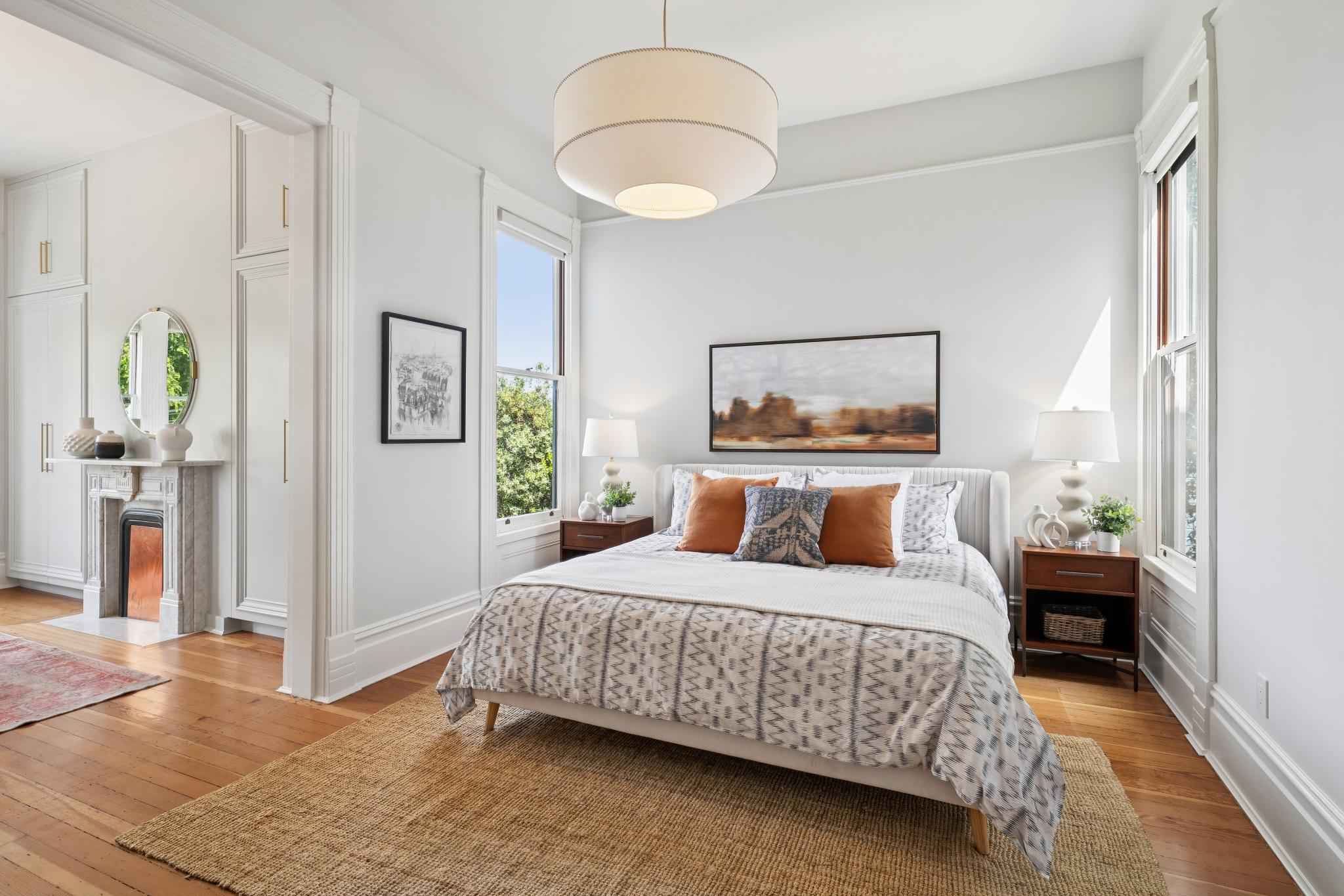 This bright and airy primary bedroom features a neutral color palette accented by warm terracotta pillows and a textured jute rug. The room is anchored by a modern upholstered bed, flanked by wooden nightstands and elegant table lamps, all illuminated by a large, contemporary drum pendant light. A fireplace with a marble surround and a circular mirror adds a touch of classic architectural charm to the adjacent space, creating a sophisticated and inviting atmosphere.