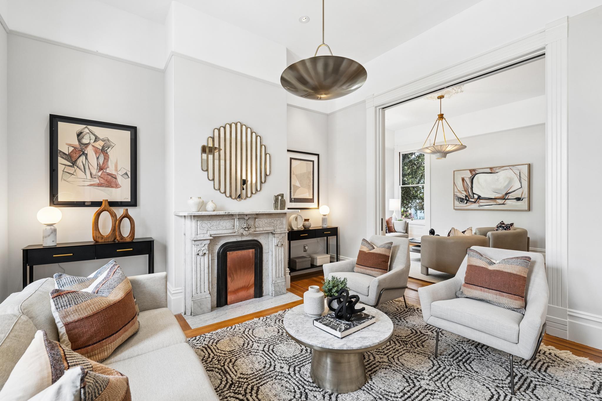 This elegant living room features a sophisticated blend of modern and classic design, anchored by a marble fireplace and a striking sunburst mirror. The space is furnished with plush, light-toned seating, a patterned area rug, and a central marble coffee table, all illuminated by a large, sculptural pendant light. The open layout flows seamlessly into an adjacent room, creating a bright and airy atmosphere perfect for refined entertaining.