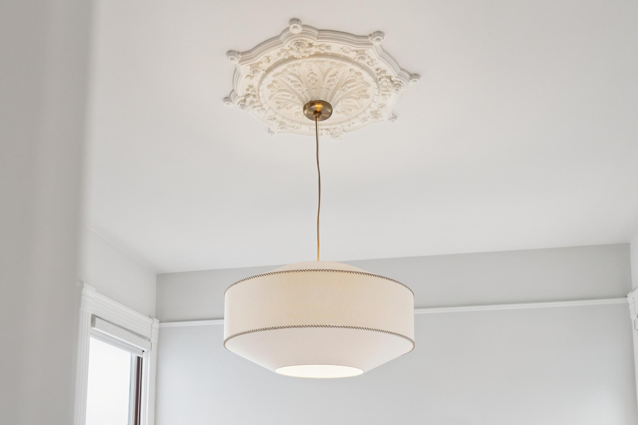 This image captures a sophisticated pendant light fixture suspended from a highly detailed, ornate white ceiling medallion. The fixture features a modern, cream-colored drum shade with subtle stitched detailing, hanging from a sleek brass-toned stem. The contrast between the classical architectural molding and the contemporary lighting design creates an elegant and refined aesthetic.