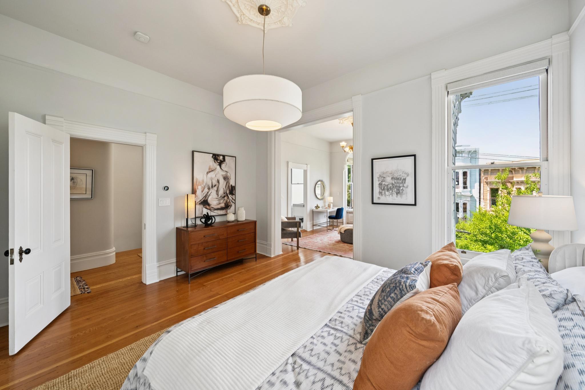 This bright and airy primary bedroom features warm hardwood floors, a large bed with layered neutral-toned bedding, and a mid-century modern wooden dresser. The room flows seamlessly into an adjacent office space through a wide doorway, while a large window provides natural light and a view of the neighborhood. The space is elegantly decorated with tasteful artwork and a classic ceiling medallion, creating a sophisticated and inviting atmosphere.