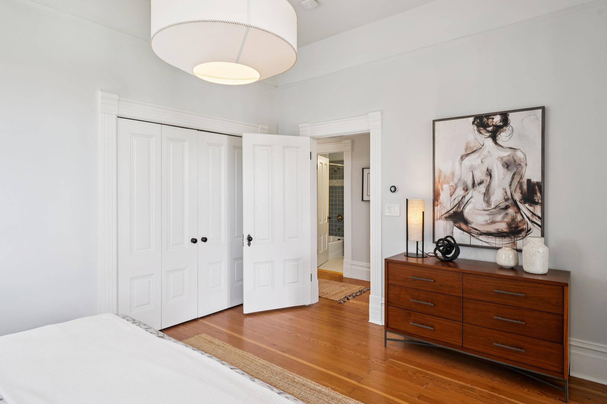 This bright and airy bedroom features warm hardwood flooring and a mid-century modern wooden dresser topped with decorative art and a sleek lamp. A large white drum pendant light hangs from the ceiling, while a set of white bi-fold closet doors and an open doorway lead to an adjacent bathroom. The room exudes a clean, minimalist aesthetic with a neutral color palette that emphasizes natural light and architectural character.